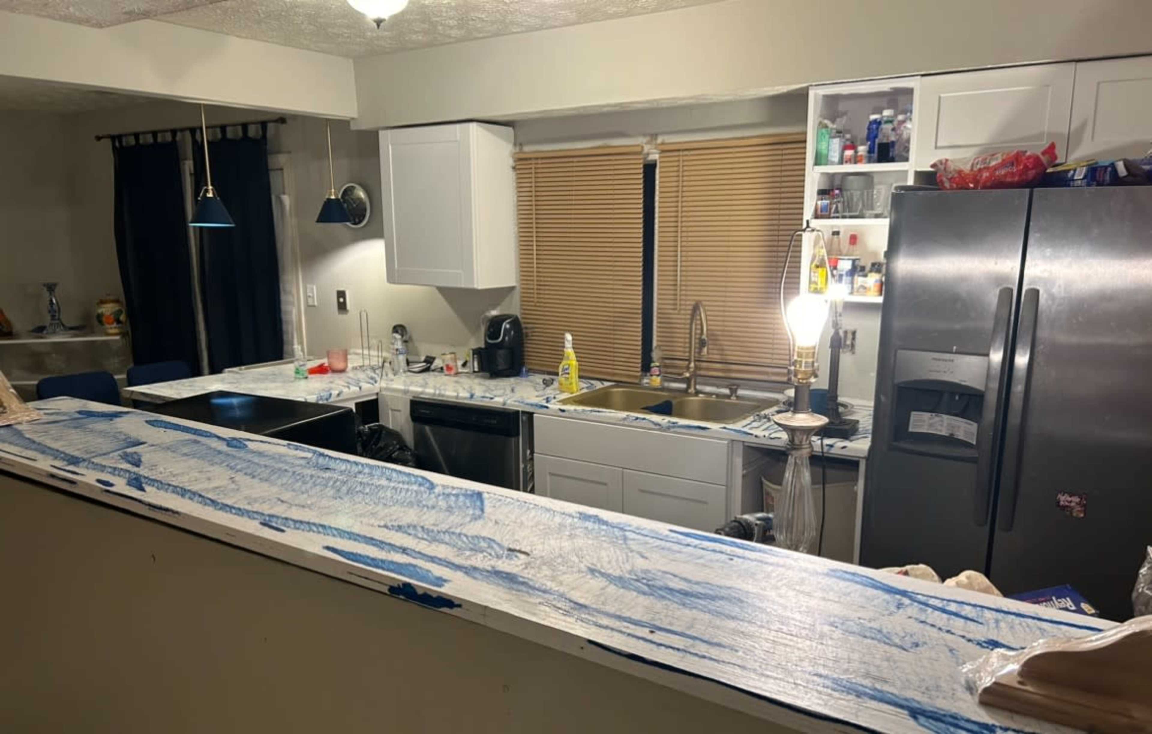 A partially renovated kitchen with a cluttered countertop, cabinets, and appliances, including a refrigerator and a sink beneath natural light from a nearby window.