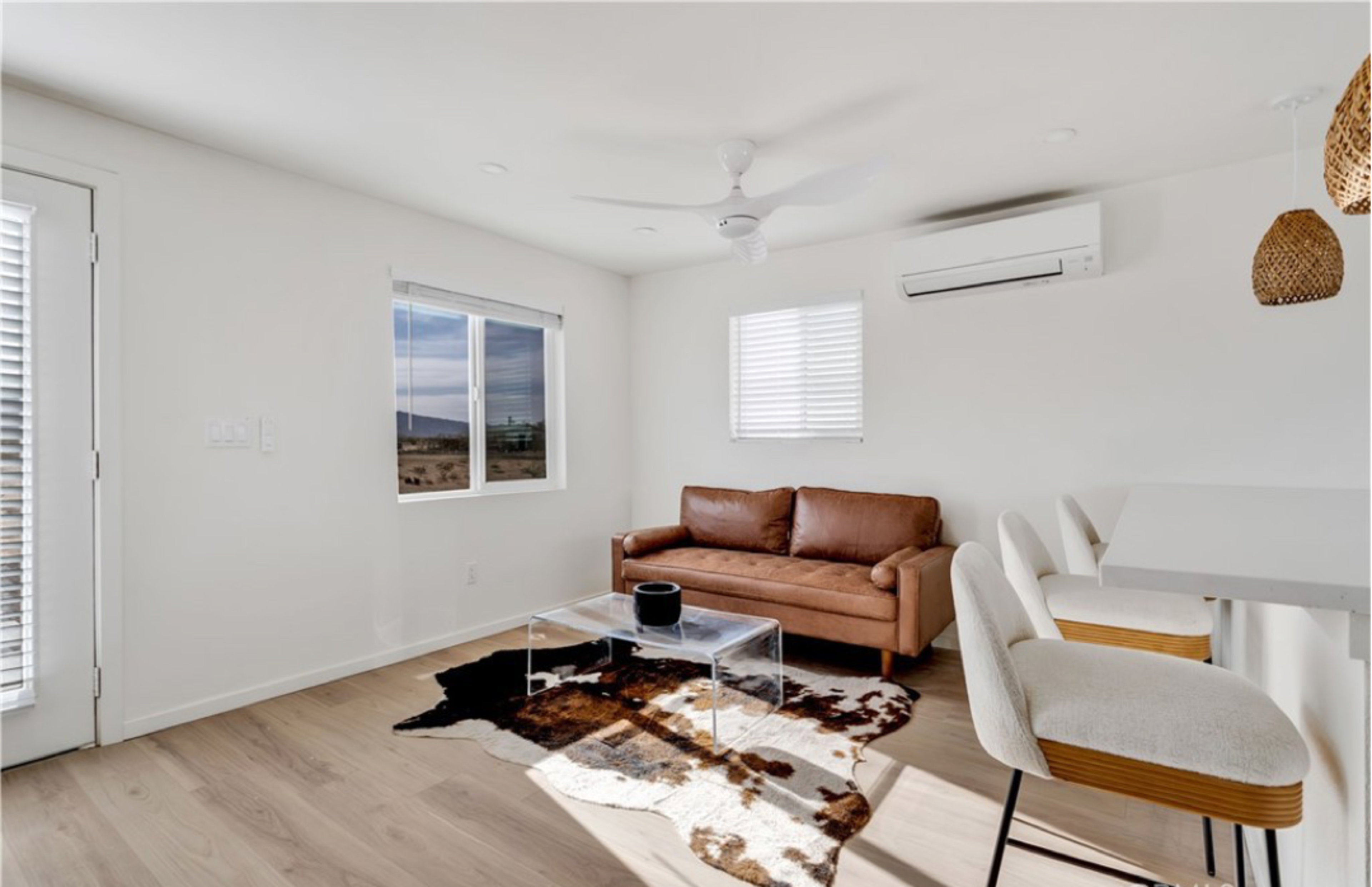 A modern, minimalist living room featuring a brown leather sofa, a clear glass coffee table, and a cowhide rug on a light wood floor.