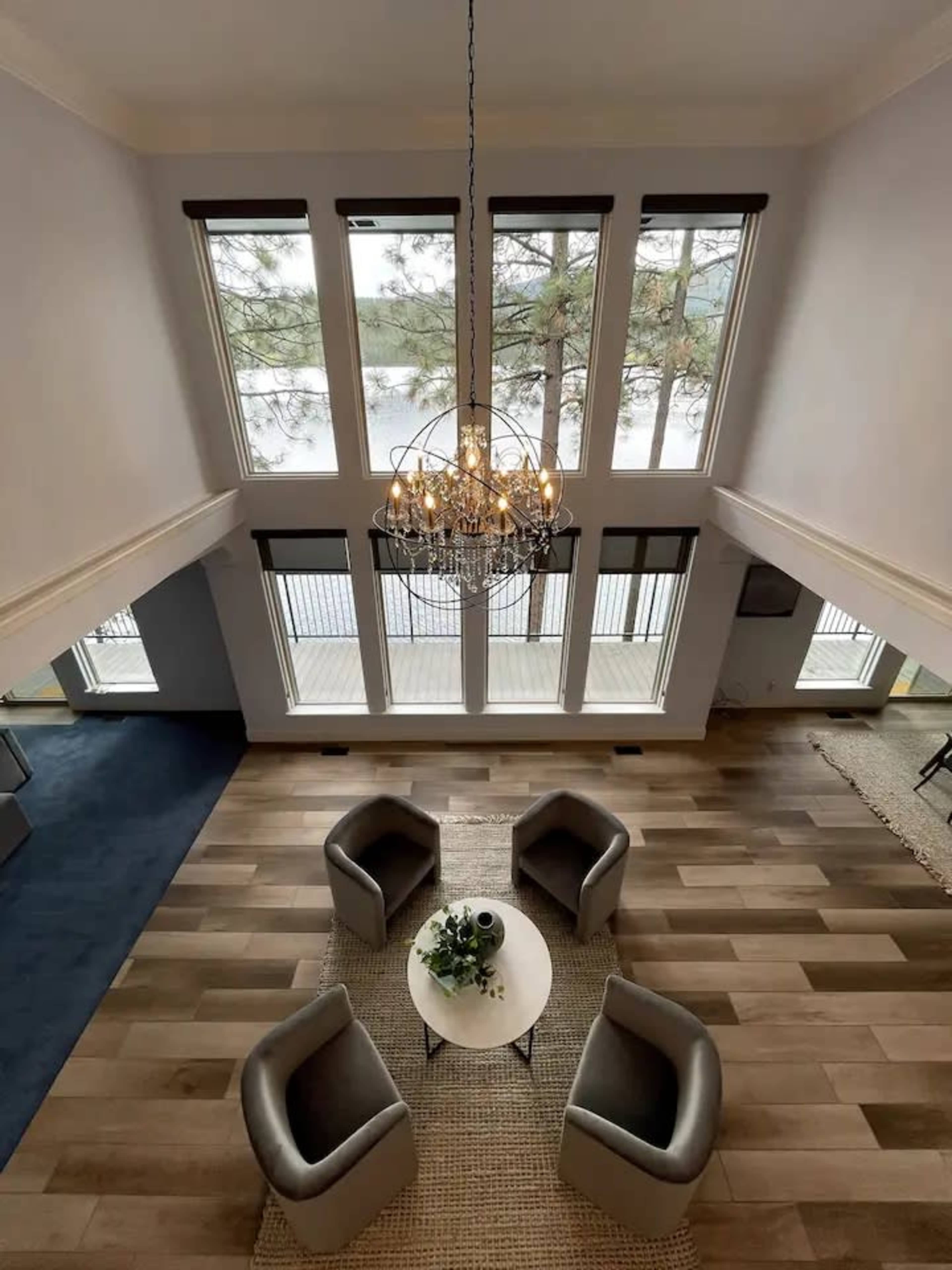 A spacious living room features four armchairs arranged around a small table, illuminated by a chandelier, with large windows overlooking a body of water.
