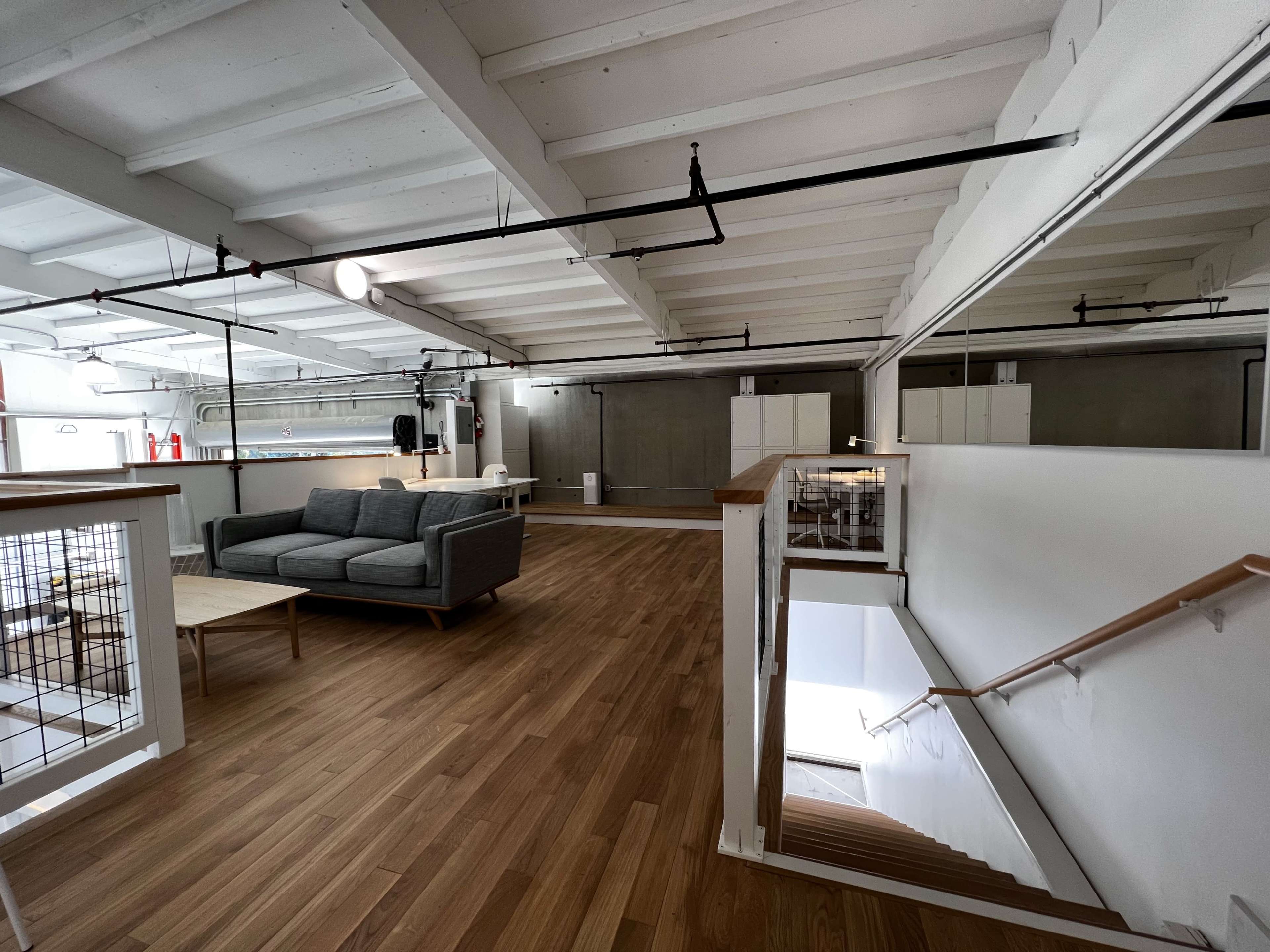 The image shows a spacious, open floor plan with wooden flooring, featuring a gray sofa and a staircase leading to another level.