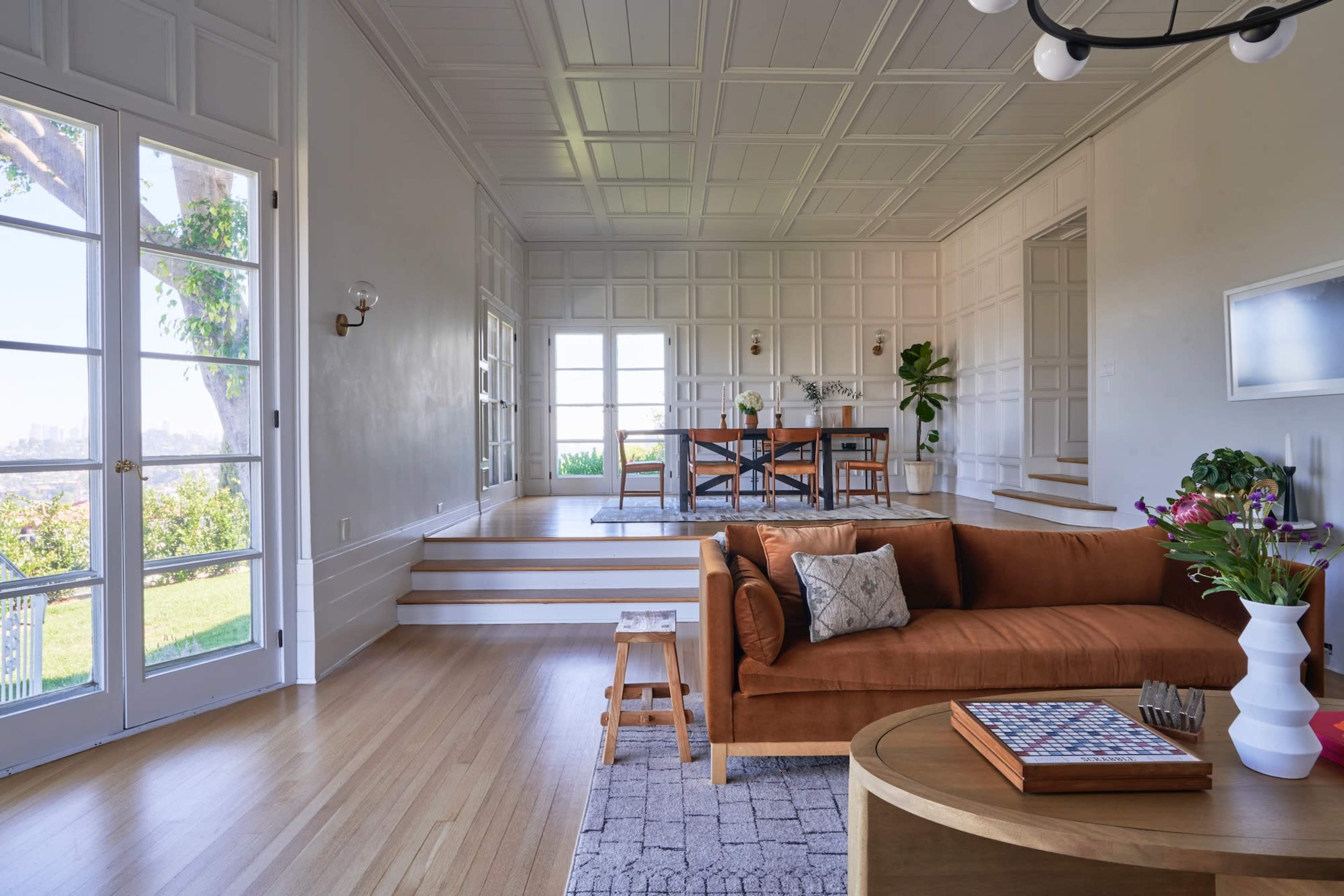 The image features a bright, open living space with a brown sofa, a round table, and large windows that provide a view of the outdoors.