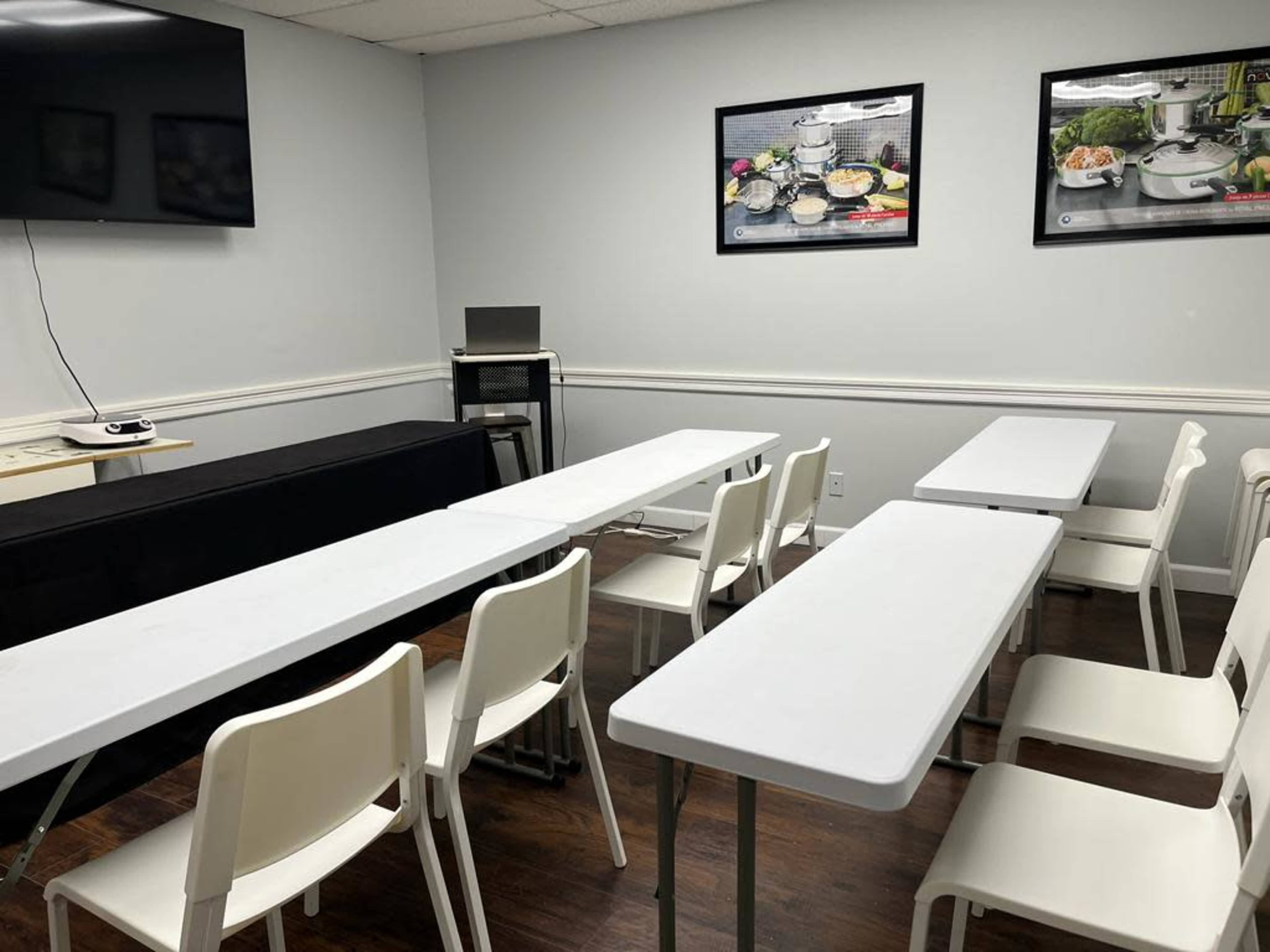A classroom or meeting space with several white tables arranged in rows and a few chairs, complemented by a television and artwork on the walls.