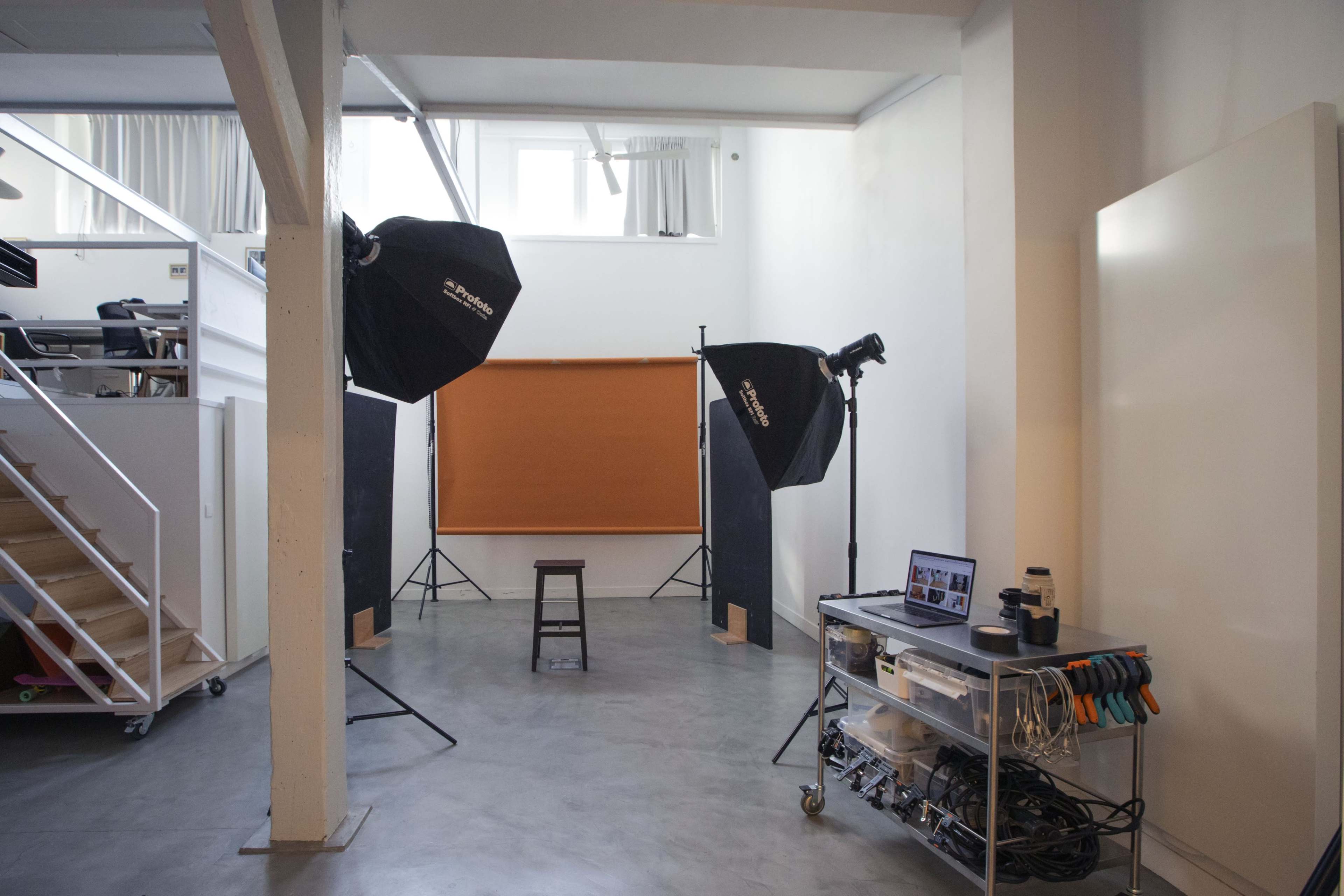 A photography studio contains two large softboxes, a backdrop, a stool, and various equipment on a rolling cart.