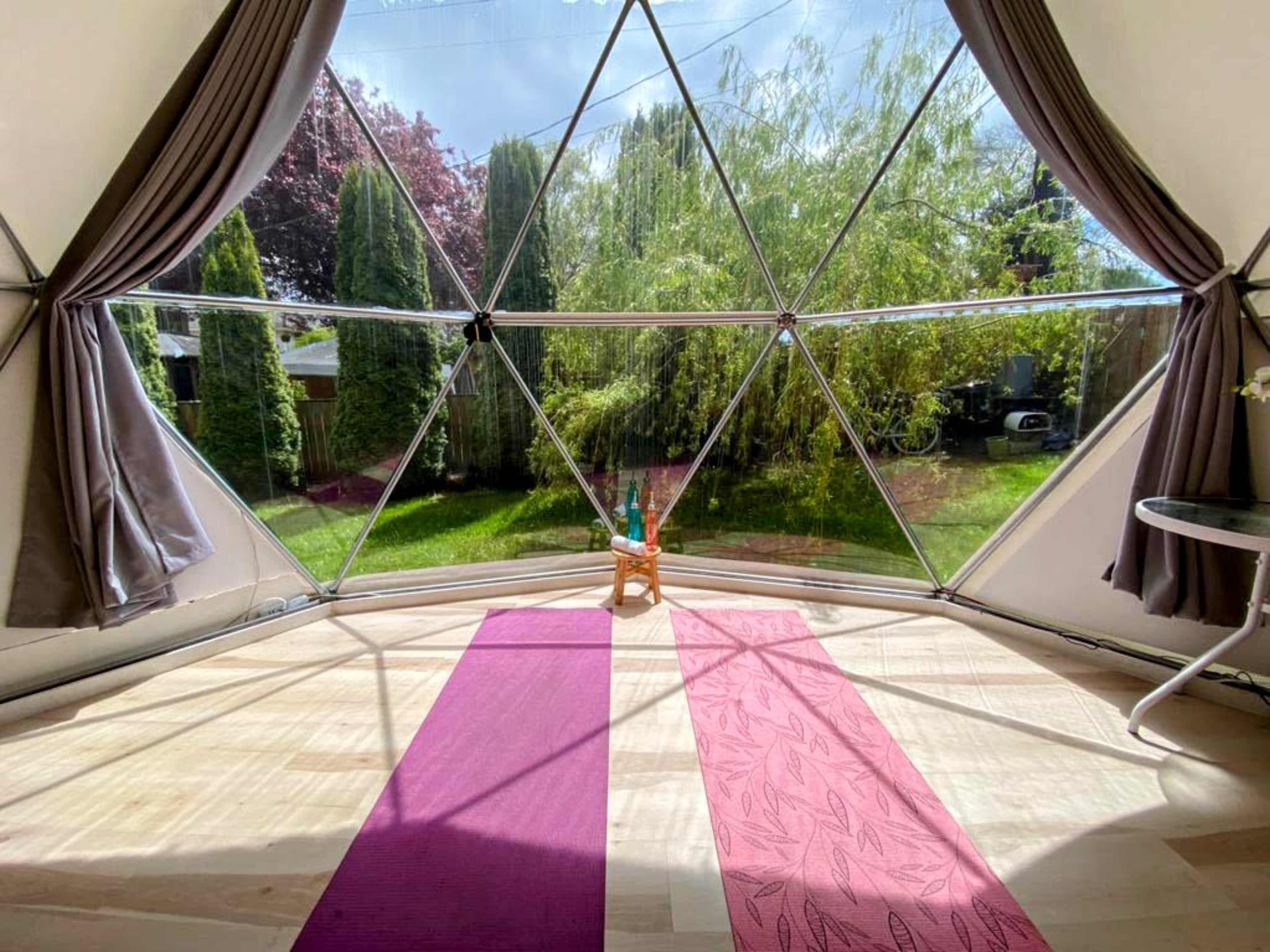 A geodesic dome interior features two yoga mats placed on the wooden floor, with large triangular windows offering a view of a green garden outside.