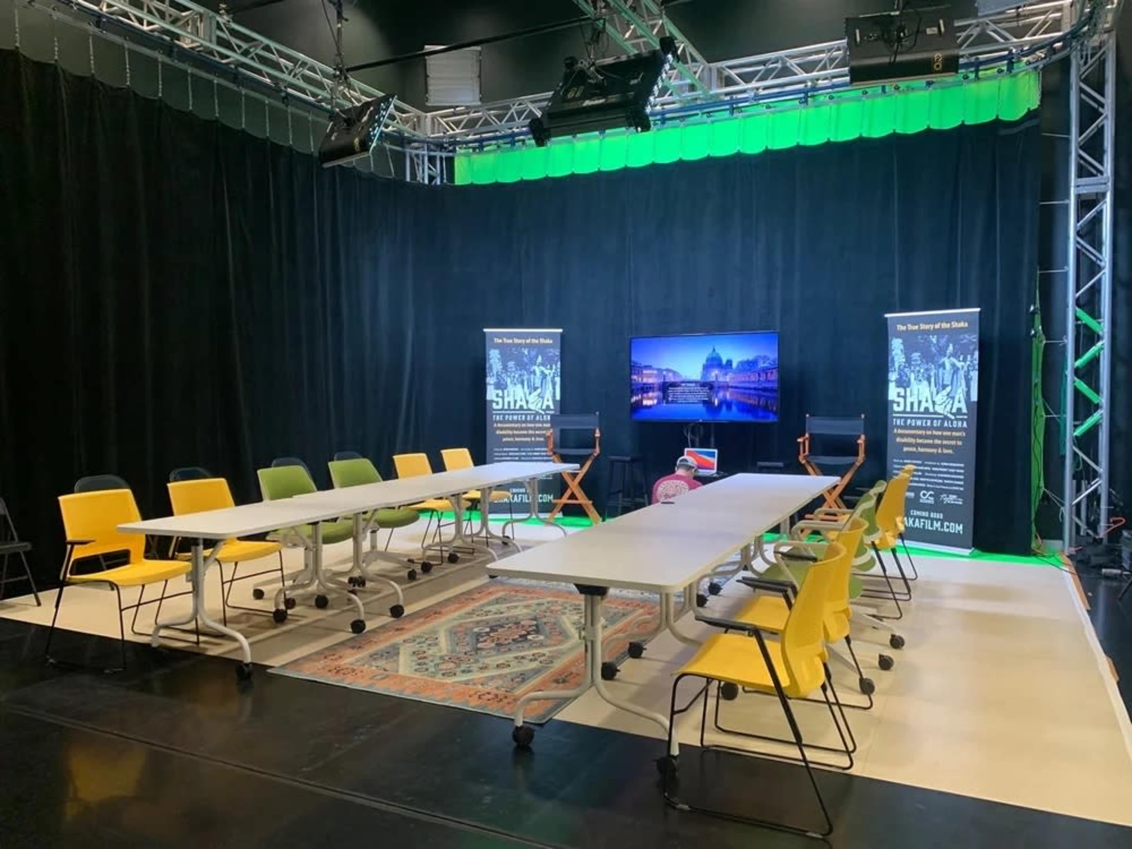 The image shows a modern studio setup with a long table surrounded by colorful chairs, a television screen displaying a cityscape, and promotional banners in the background.