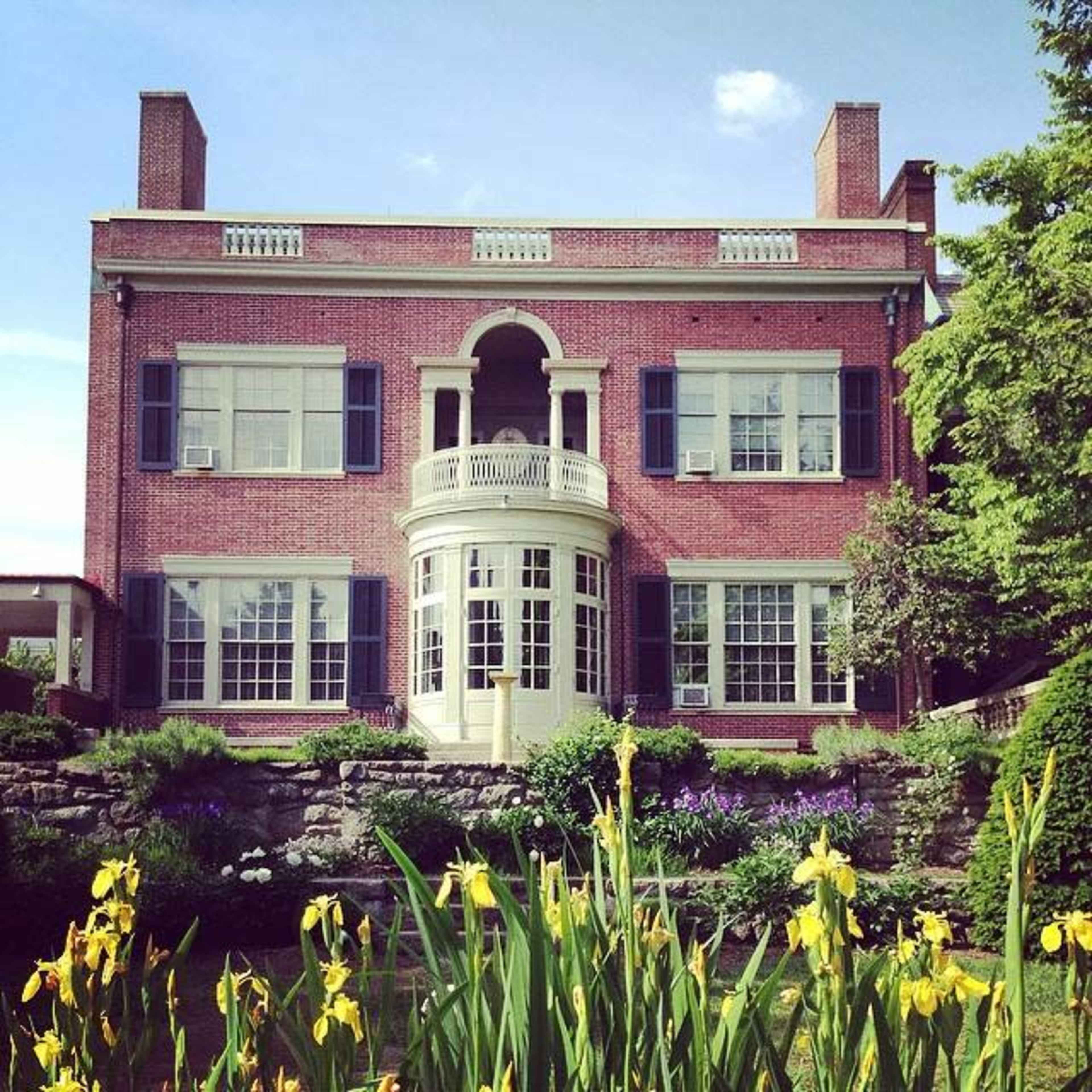 A red brick house features large windows and a central balcony, surrounded by a landscaped garden with yellow flowers.