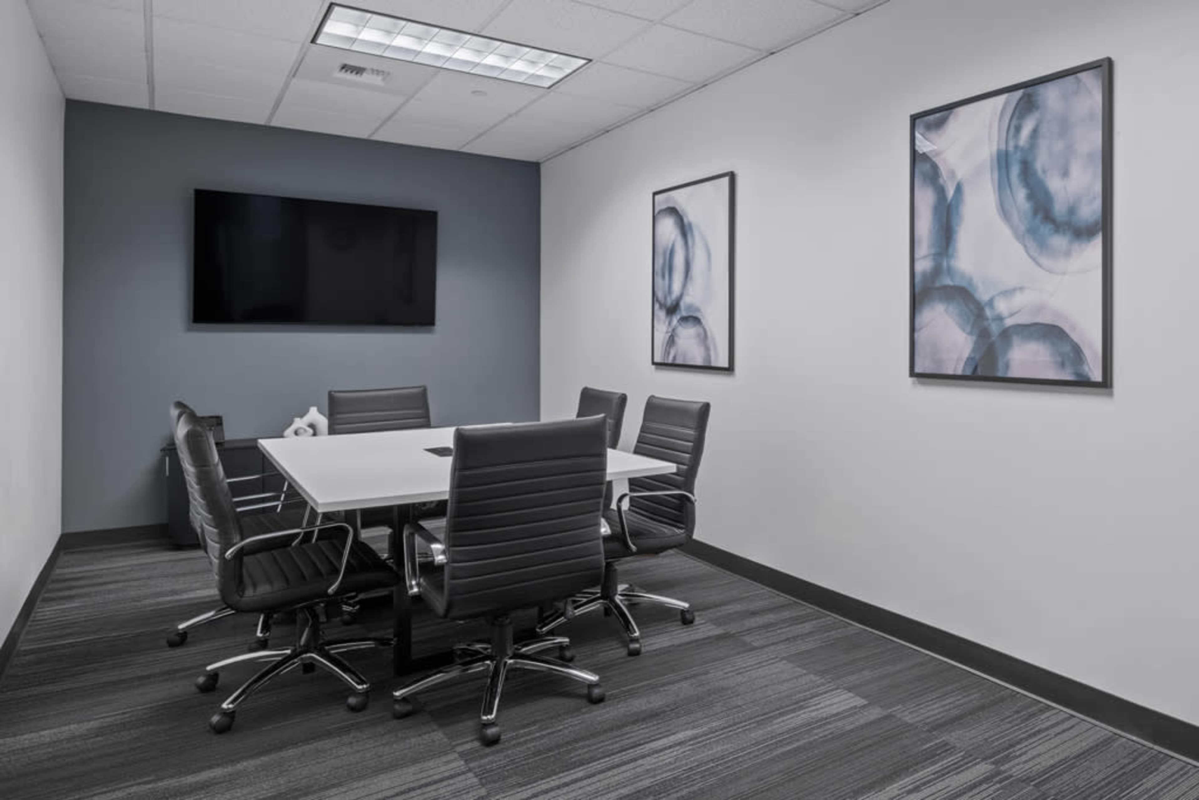 The image shows a modern conference room featuring a rectangular table surrounded by black office chairs, with two abstract artworks on the walls and a television mounted on the front wall.