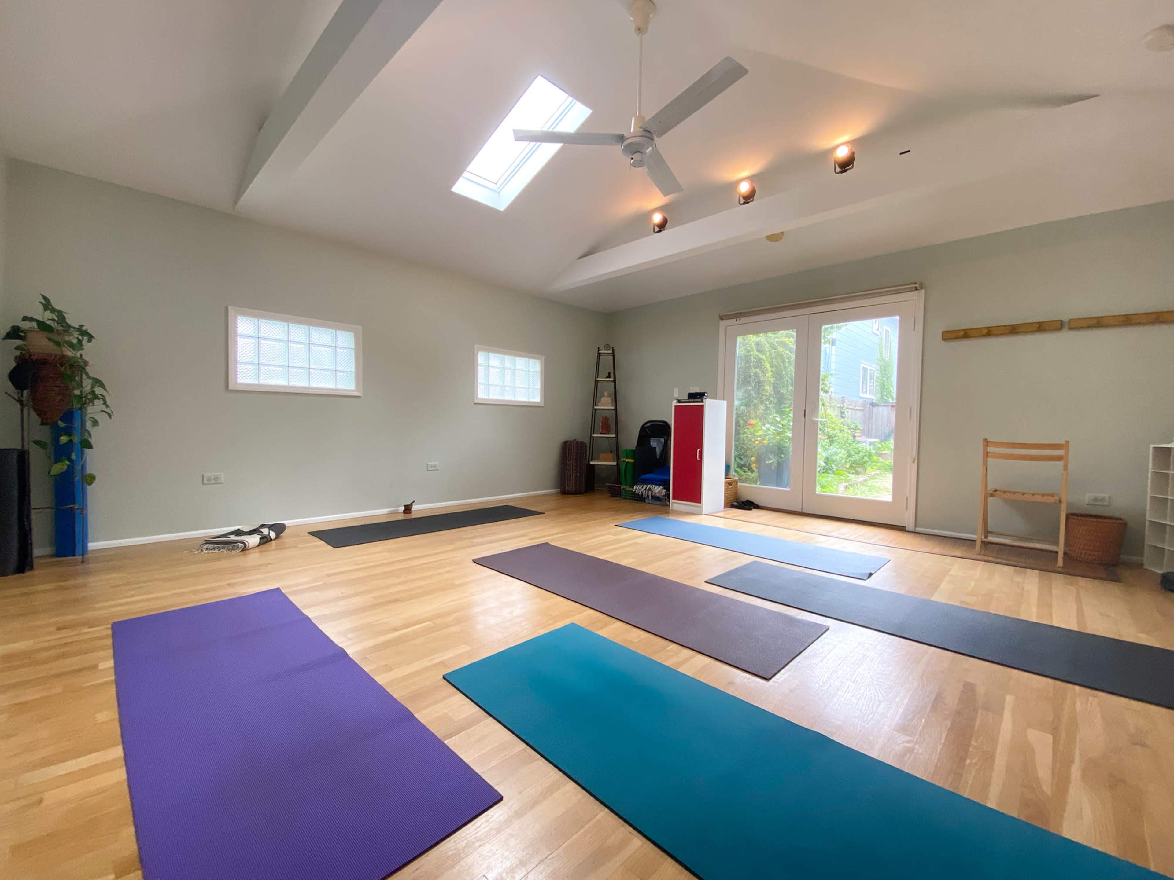 A bright, spacious yoga studio with wooden floors, multiple yoga mats arranged on the floor, and large windows letting in natural light.