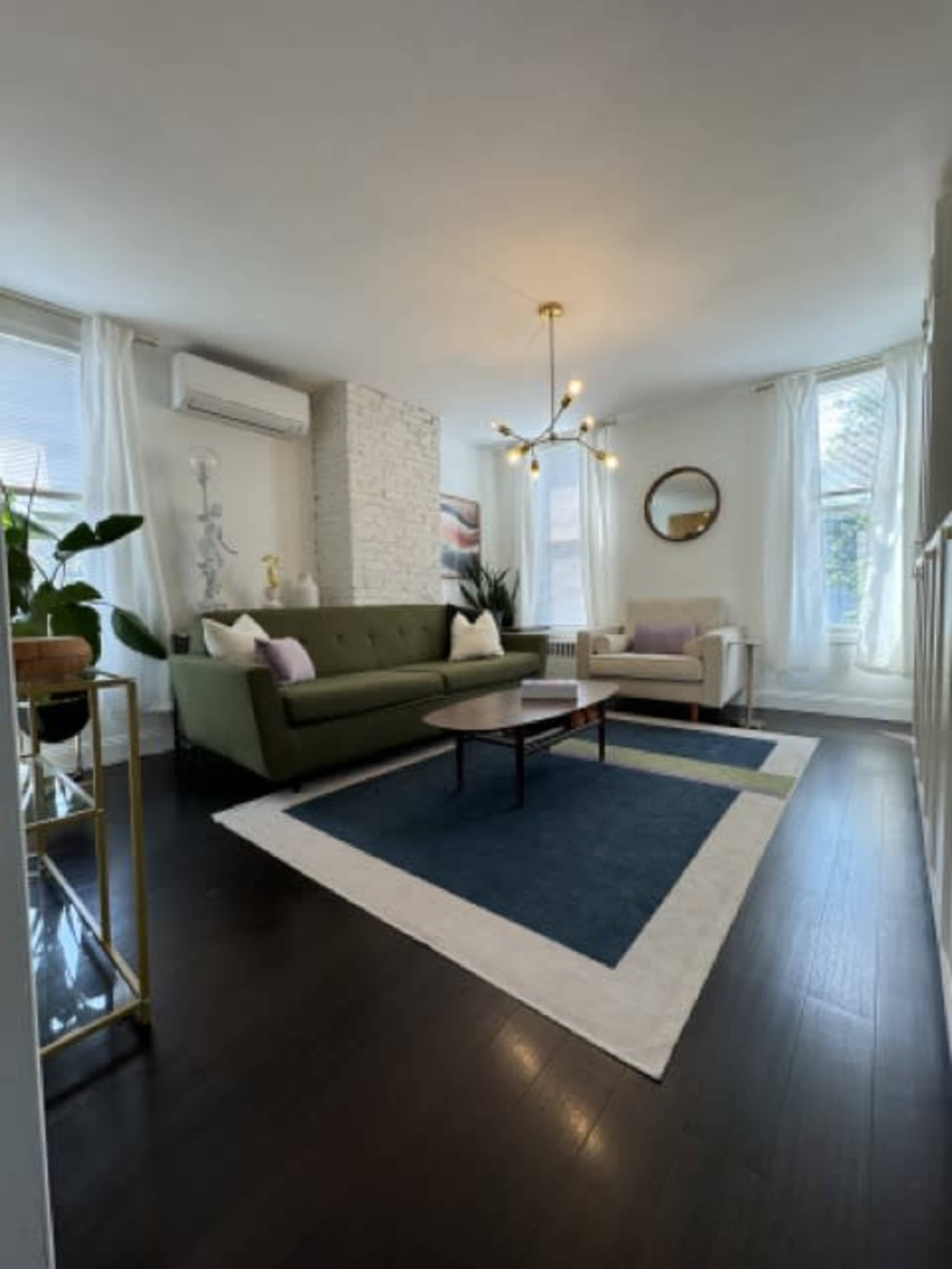 The image shows a modern living room featuring a green sofa, a white armchair, a blue area rug, and a decorative chandelier, with large windows allowing natural light to enter.