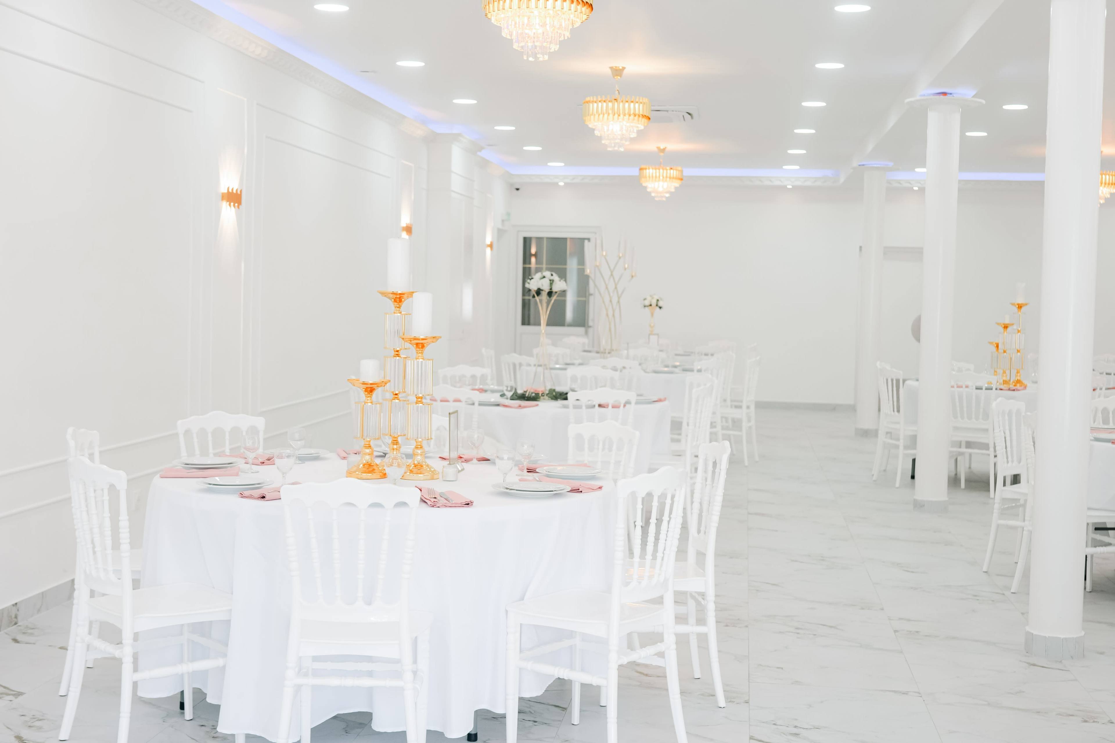 The image shows a banquet hall with white tables set for a dinner, adorned with golden candle holders and elegant tableware under soft lighting.