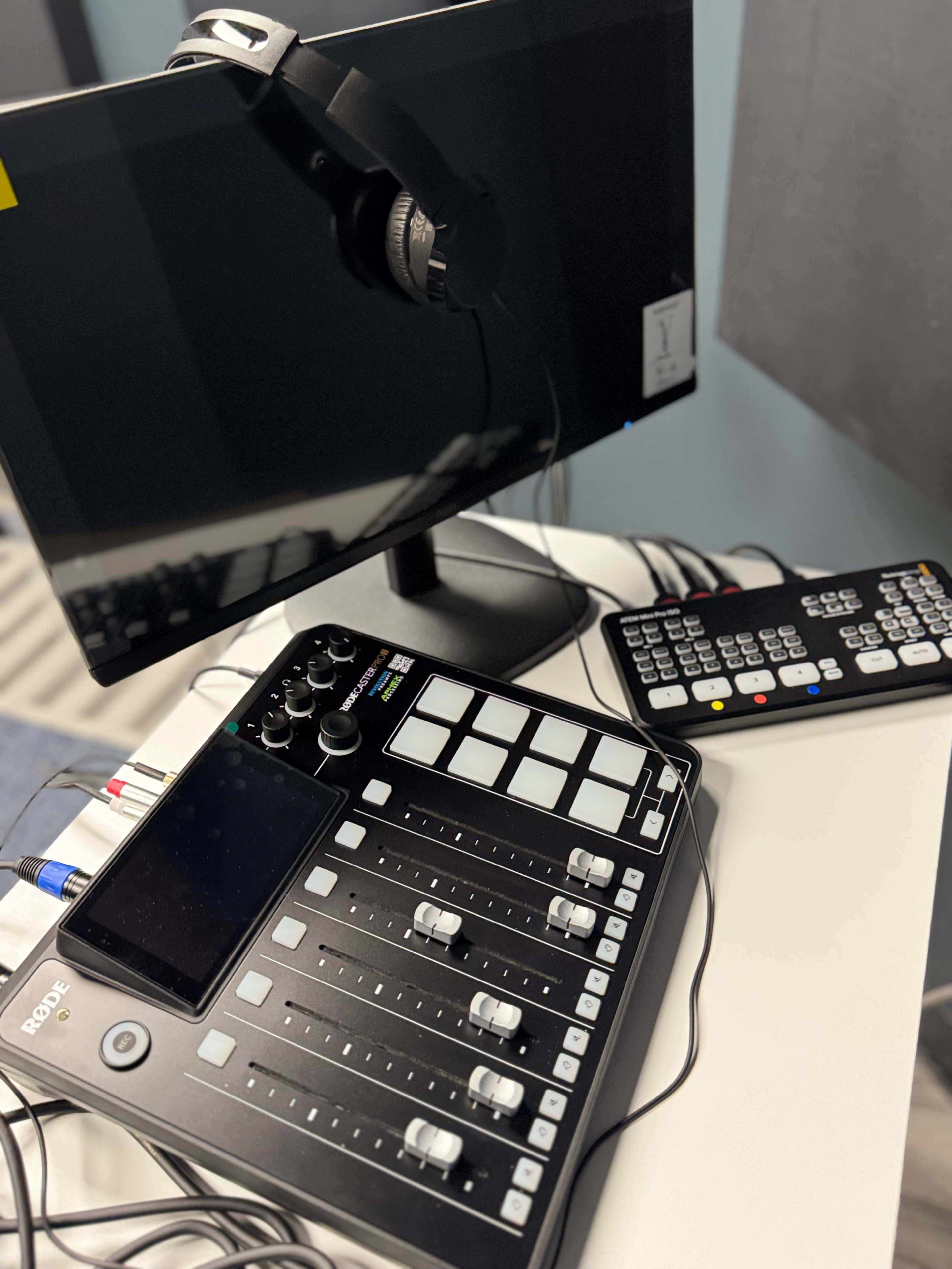 The image shows an audio mixing console with several sliders and buttons, a monitor, a pair of headphones on top, and a remote control on a table.