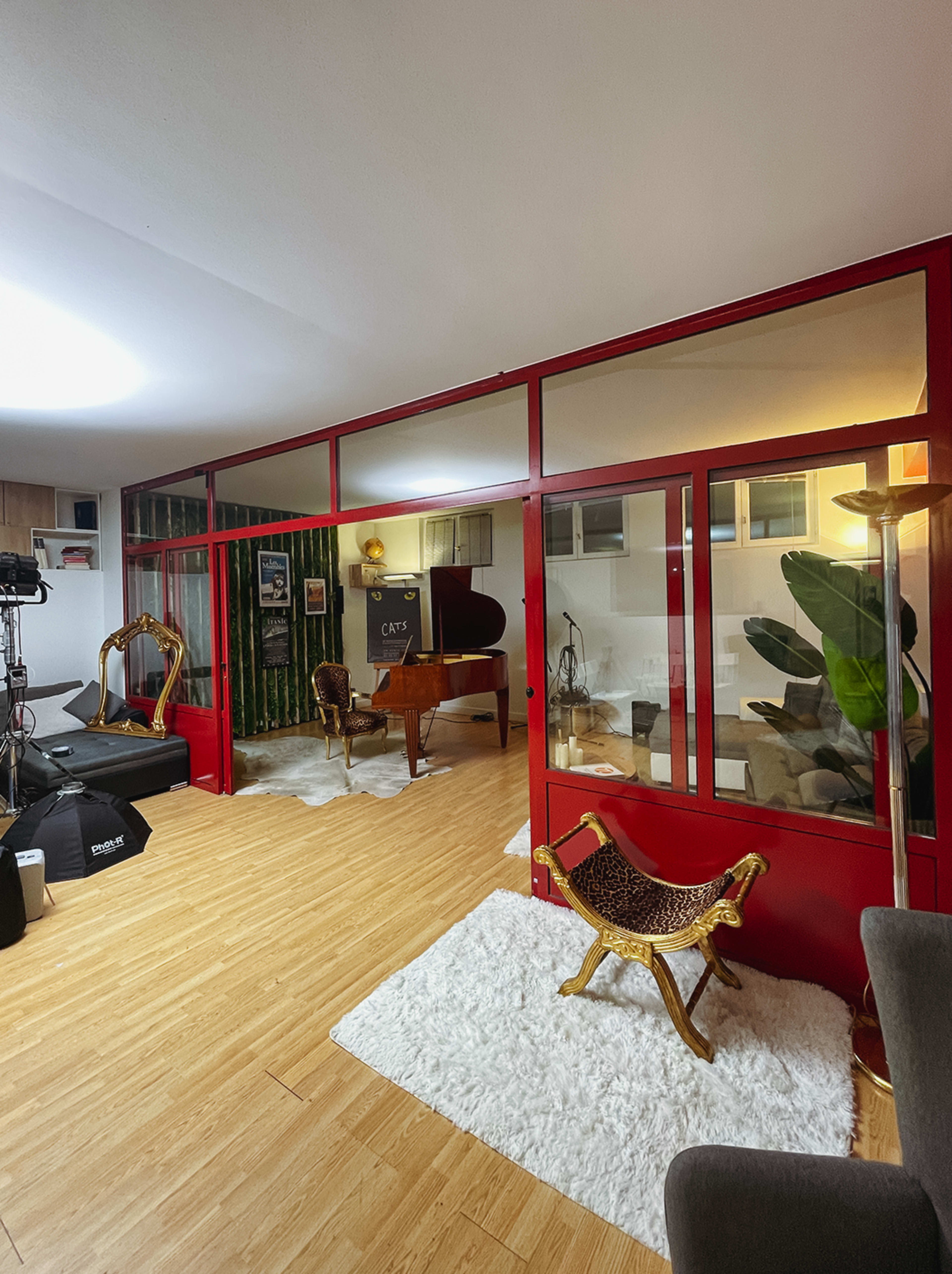 The image shows a modern studio space with hardwood flooring, featuring a grand piano, plants, and various seating arrangements in a divided area.
