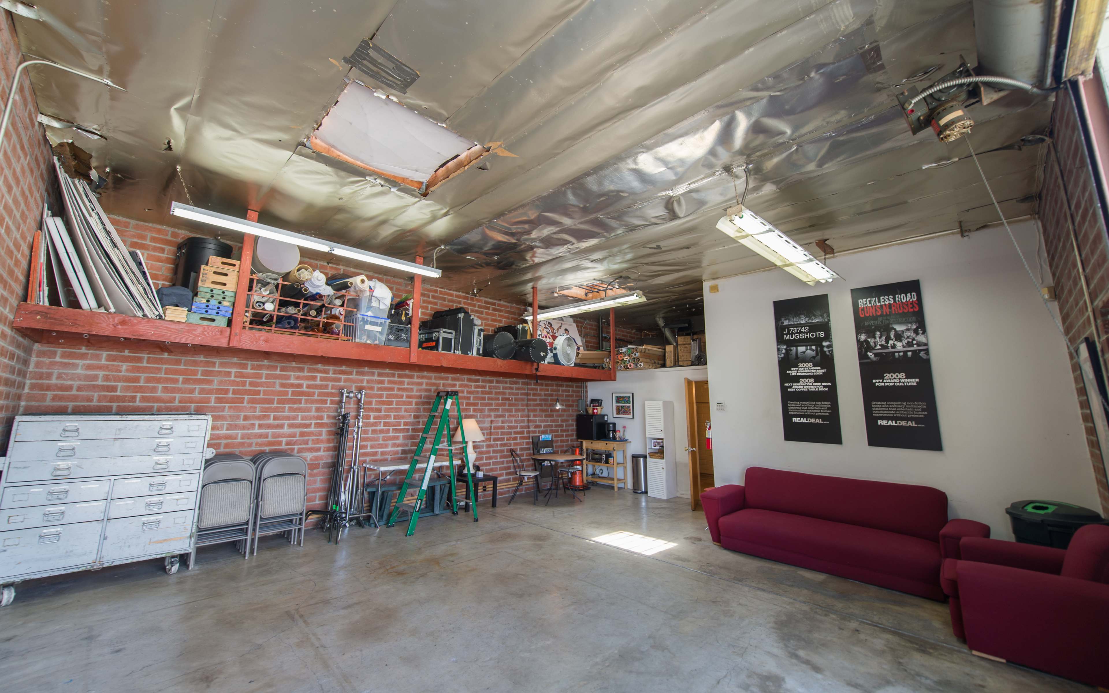 The image shows a spacious garage with bare concrete floors, exposed brick walls, a red couch, metal chairs, and a loft area containing various items.