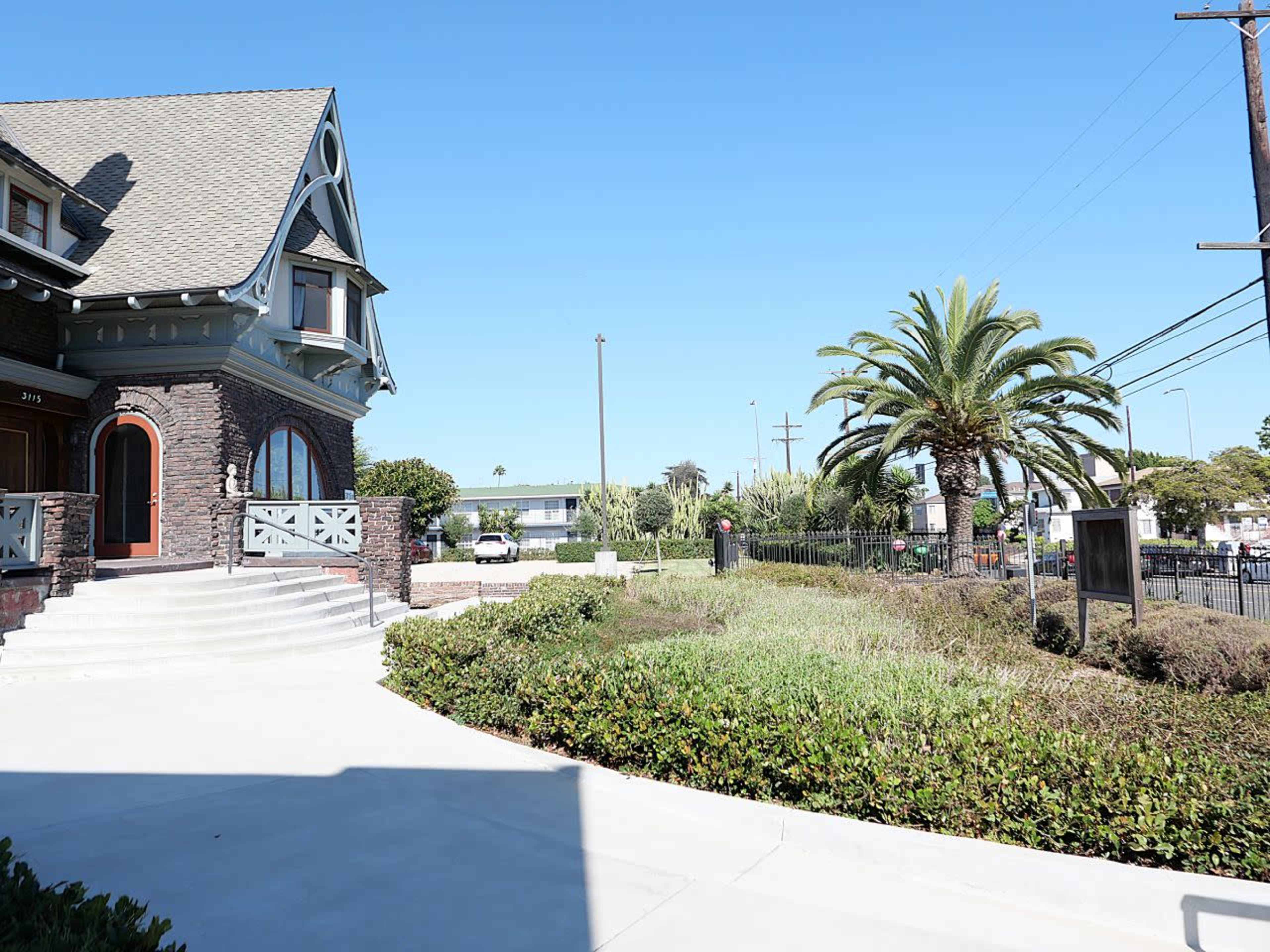 The image shows a large house with a peaked roof and decorative elements, surrounded by landscaped greenery and palm trees, under a clear blue sky.