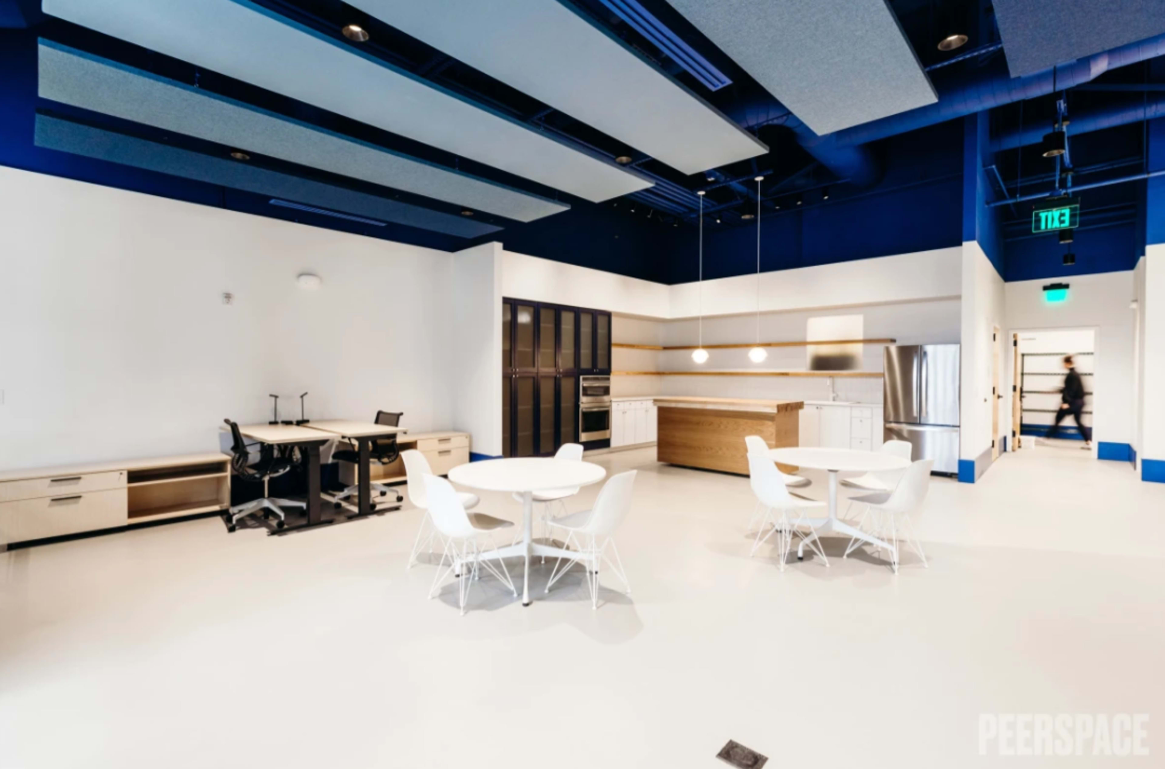 A modern office space with a kitchen area, a table with four chairs, and workstations set against a bright, minimalist backdrop.