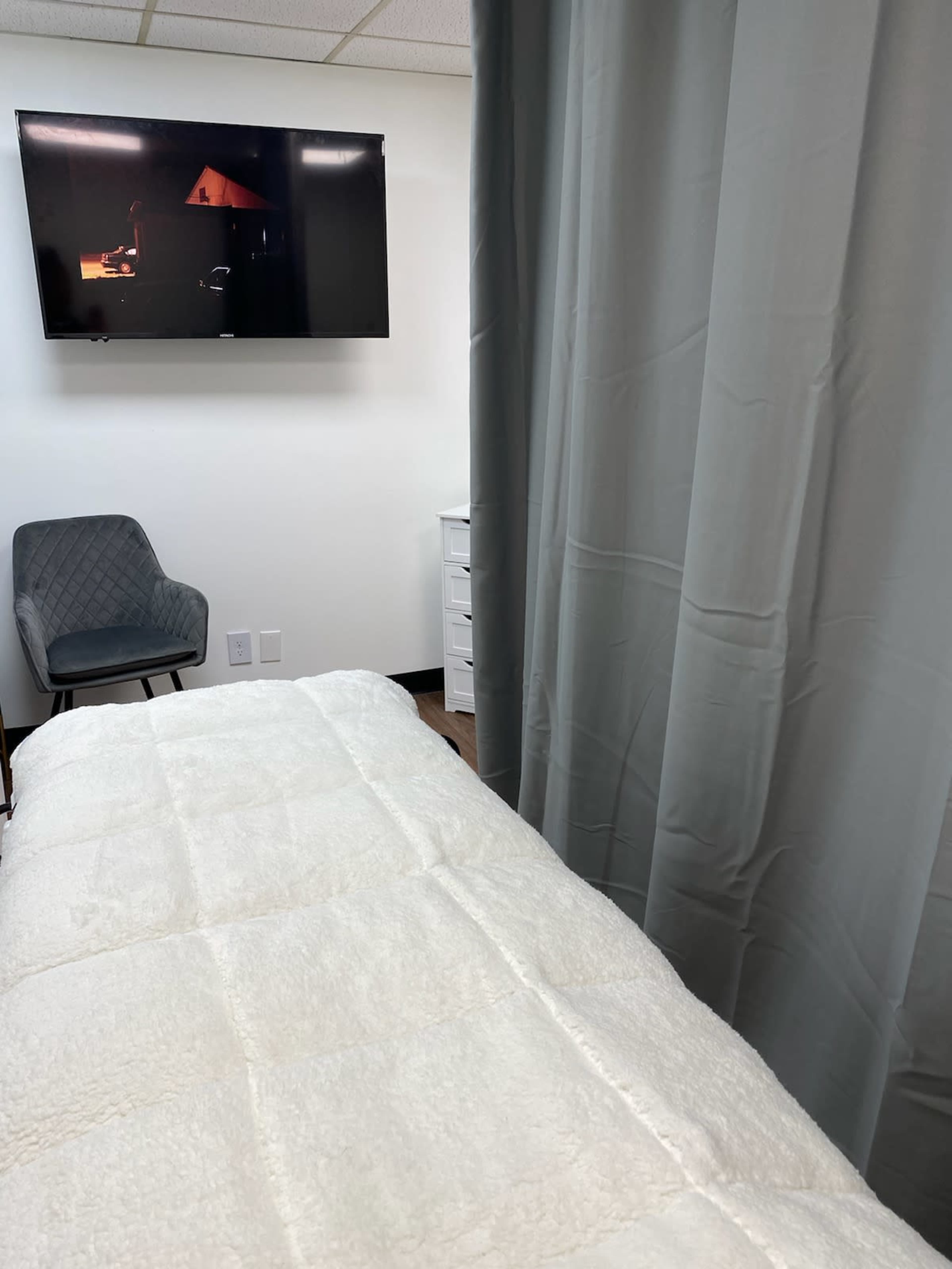 The image shows a treatment room with a white bed covered in a soft blanket, a grey chair in the corner, and a television mounted on the wall displaying a dark scene.
