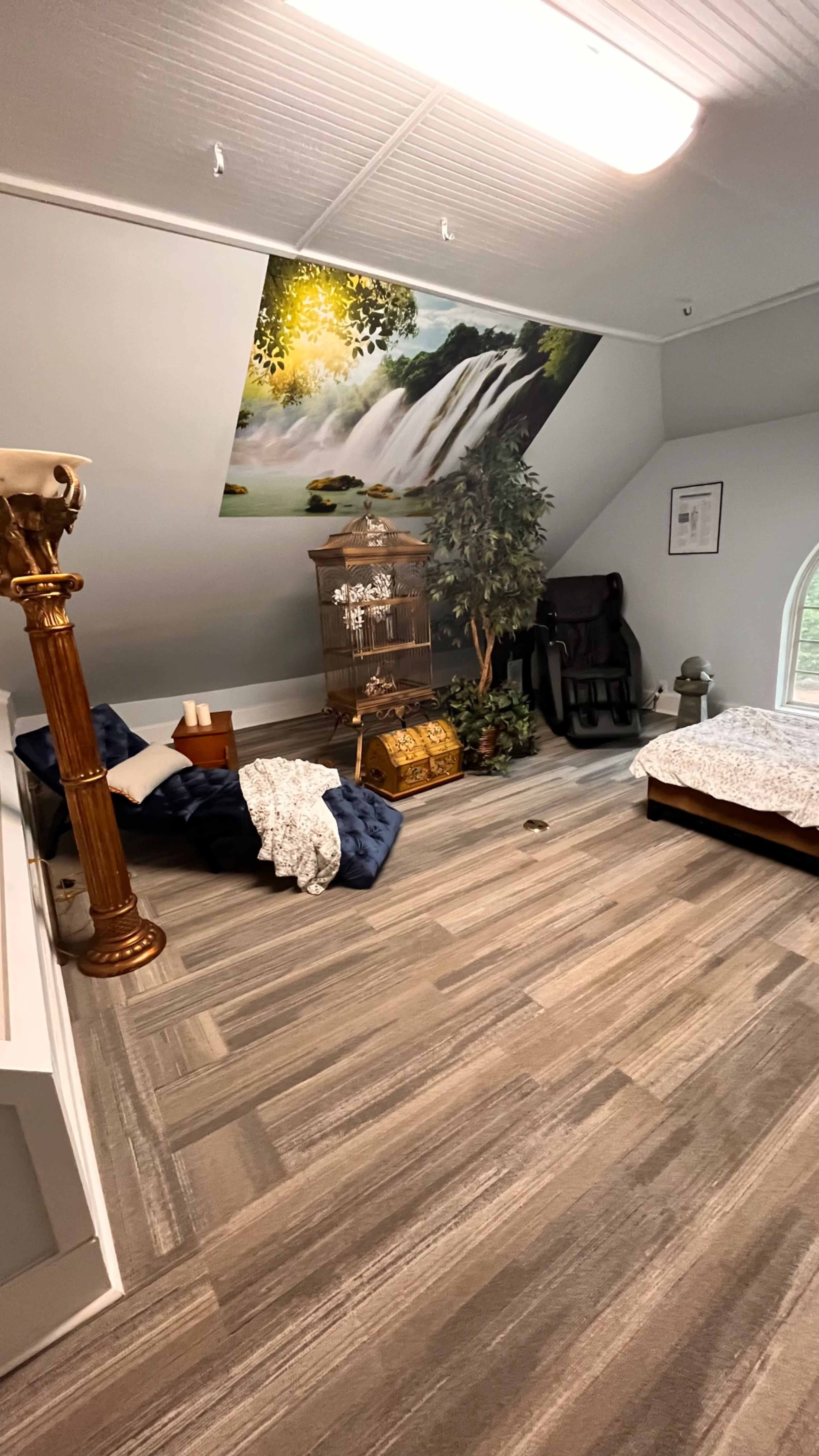 A spacious attic room featuring a bed, a vintage chair, a decorative parrot cage, and a mural of a waterfall on the wall.