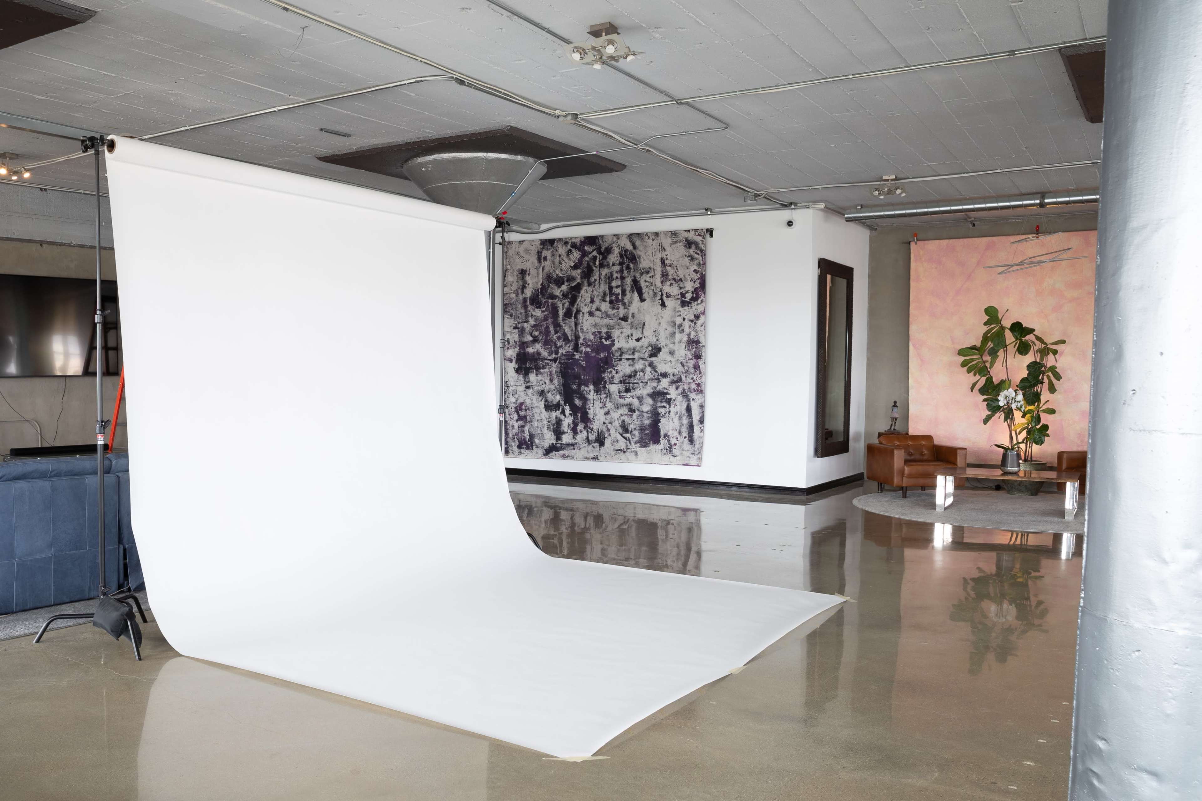 An empty studio space featuring a white backdrop, polished concrete floors, and a large piece of abstract art on the wall, along with a seating area with two leather chairs and a potted plant.