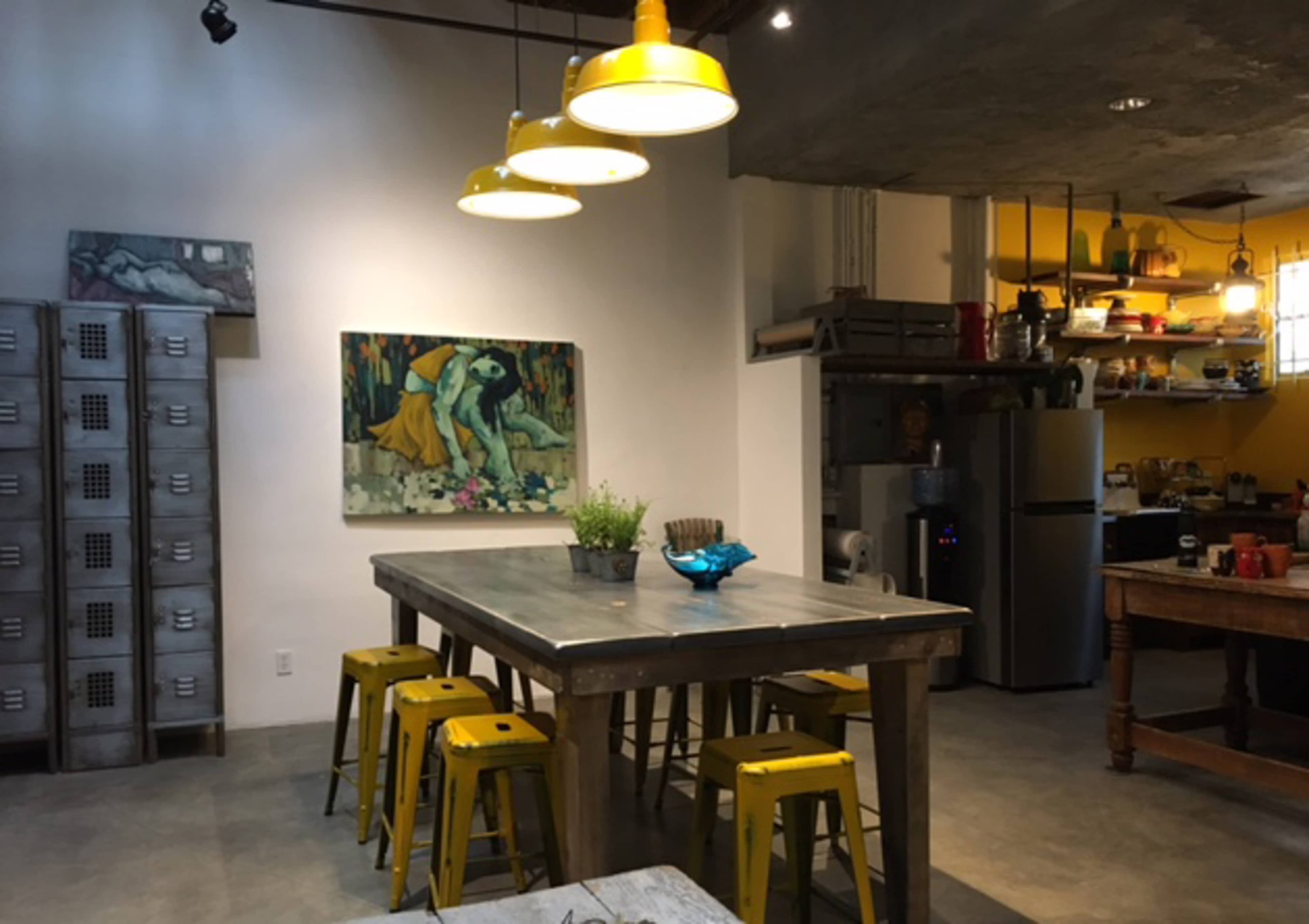A dining area features a large wooden table surrounded by yellow metal stools, illuminated by pendant lights, with a colorful painting on the wall and shelves holding various kitchen items in the background.