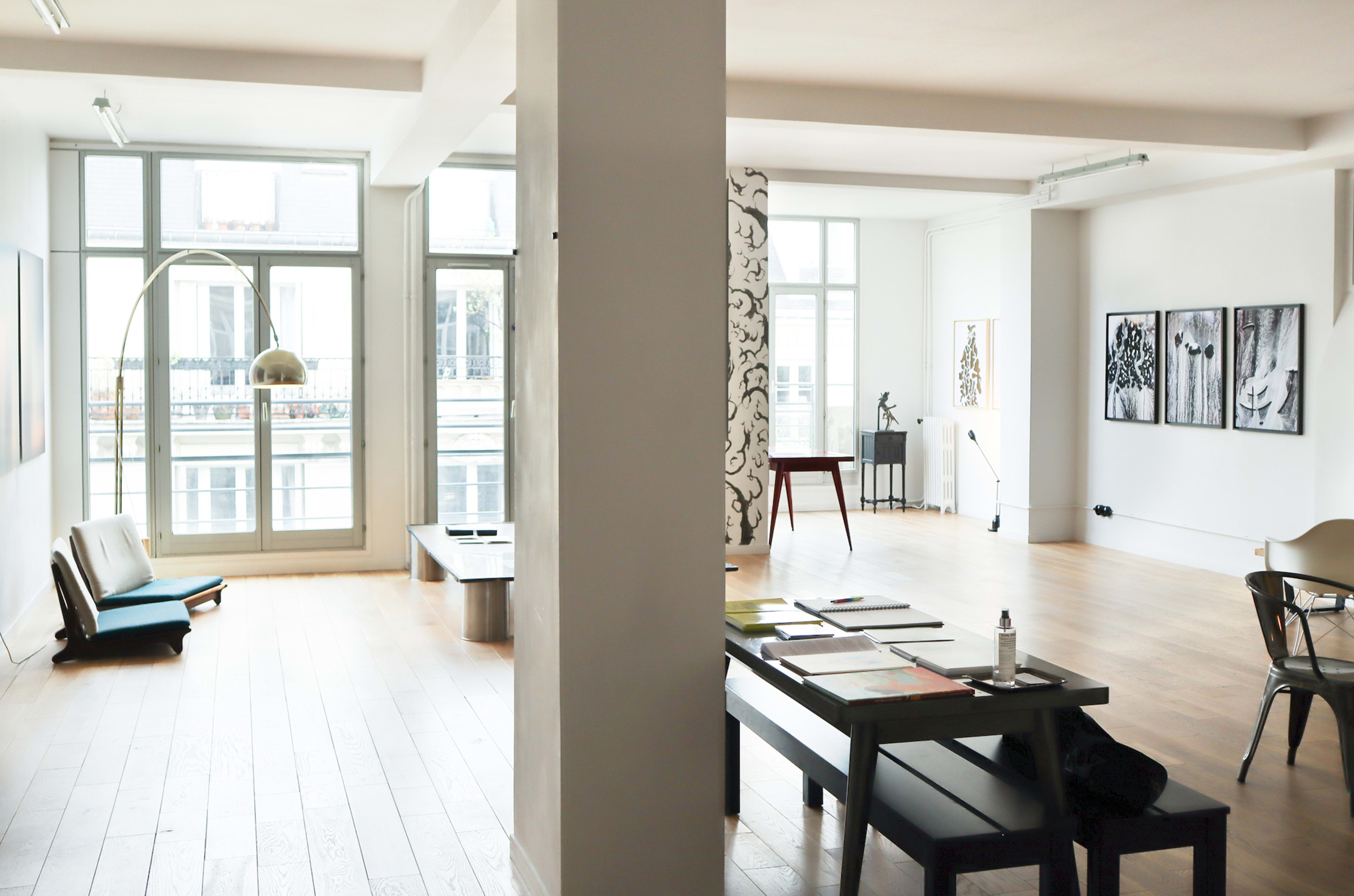The image shows a spacious, modern room with large windows, wooden flooring, and various pieces of furniture including a sofa, tables, and artwork on the walls.