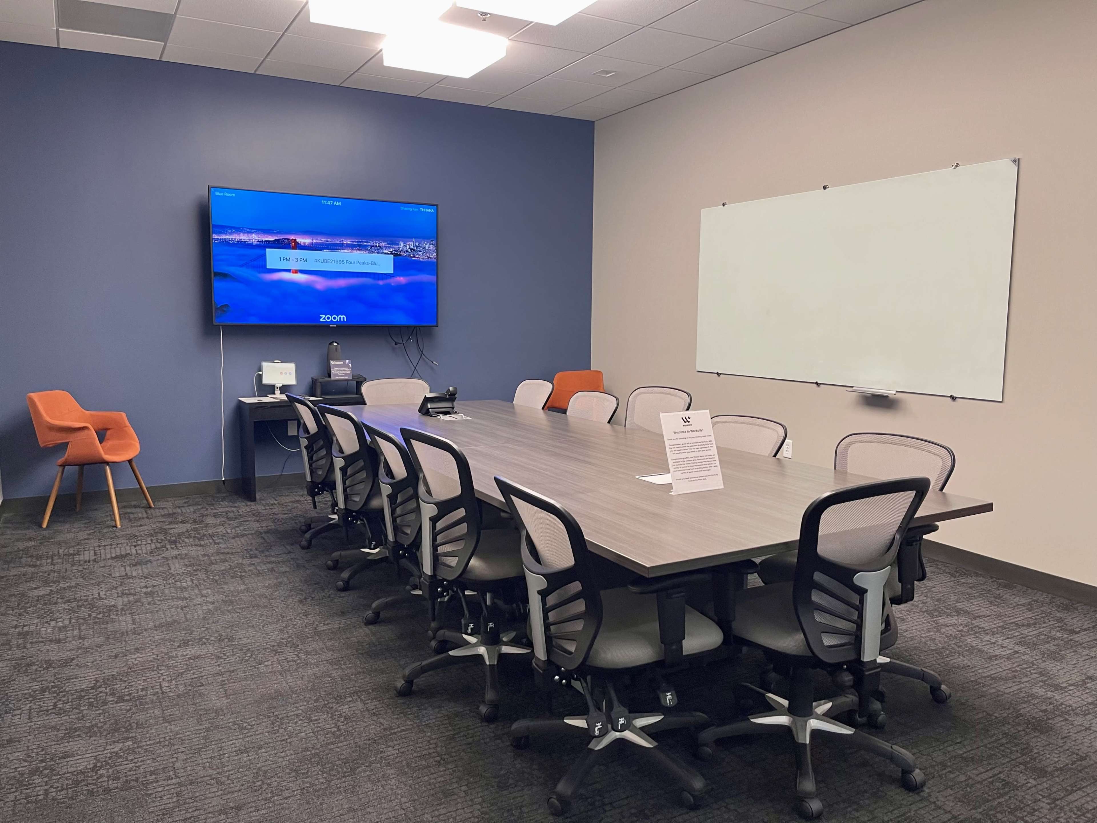 A conference room is set up with a long table surrounded by chairs, a large screen displaying a Zoom interface, and a whiteboard on the wall.