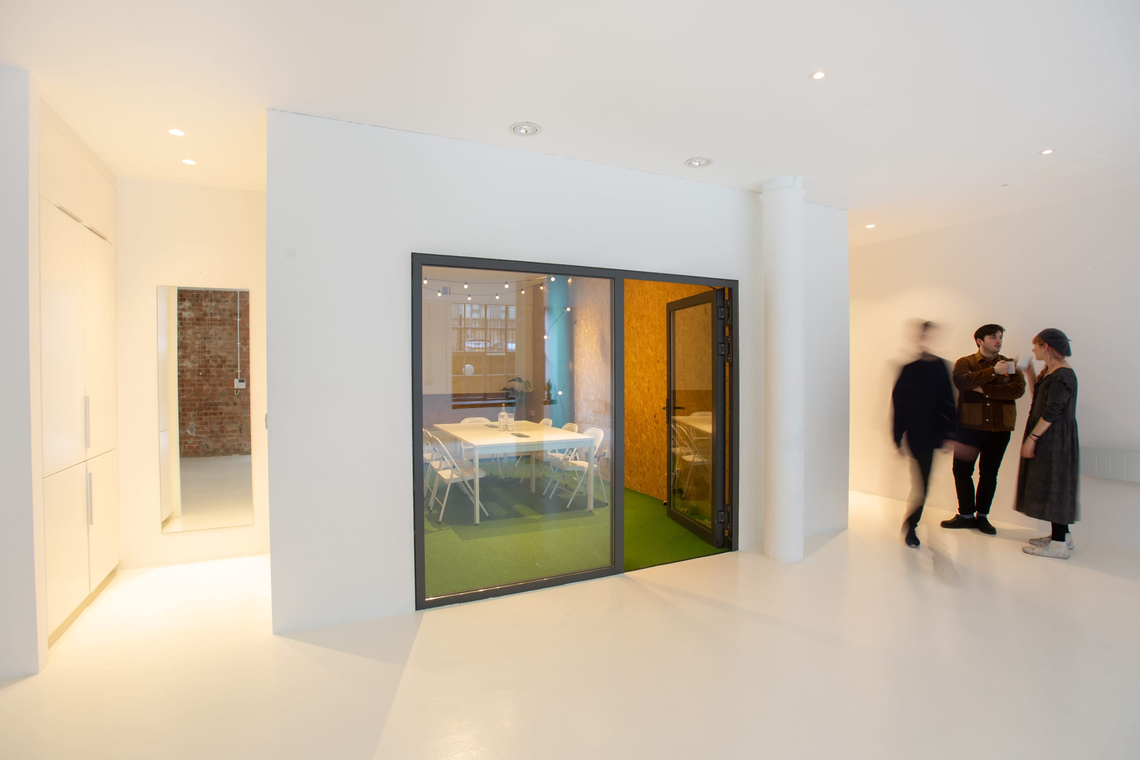 A bright, modern office space features a meeting room with glass walls, a green carpet, and three individuals engaged in conversation nearby.