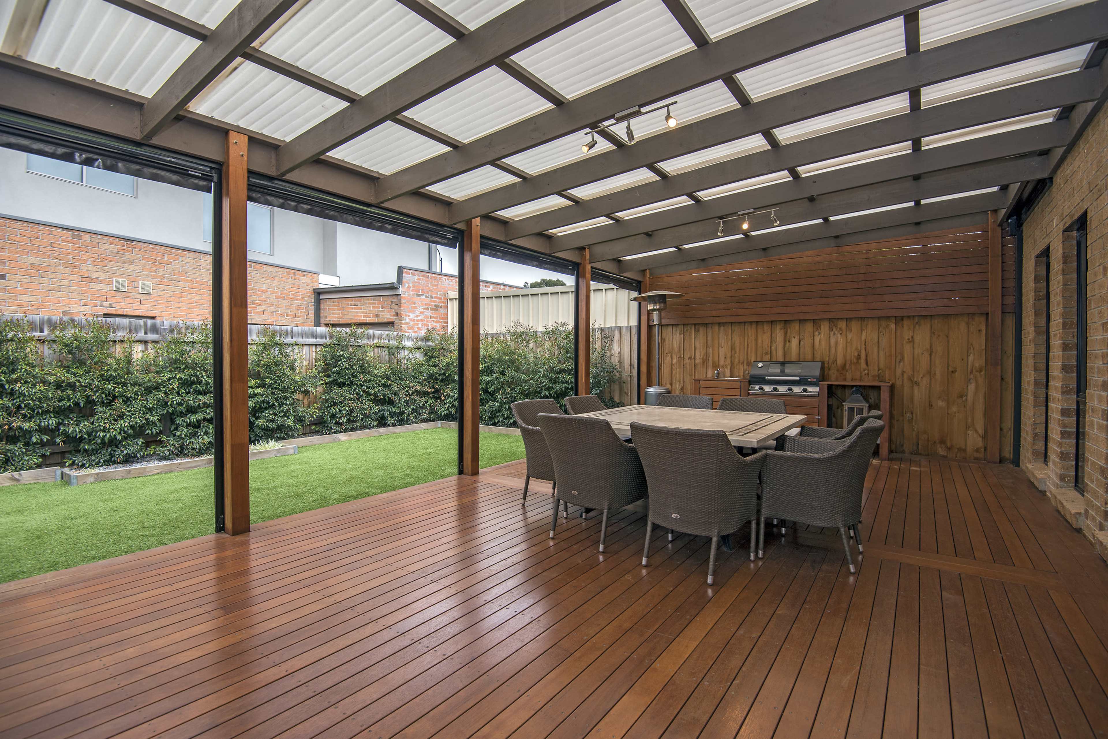 A covered outdoor patio with a dining table and chairs, a barbecue grill, and landscaped greenery in the background.