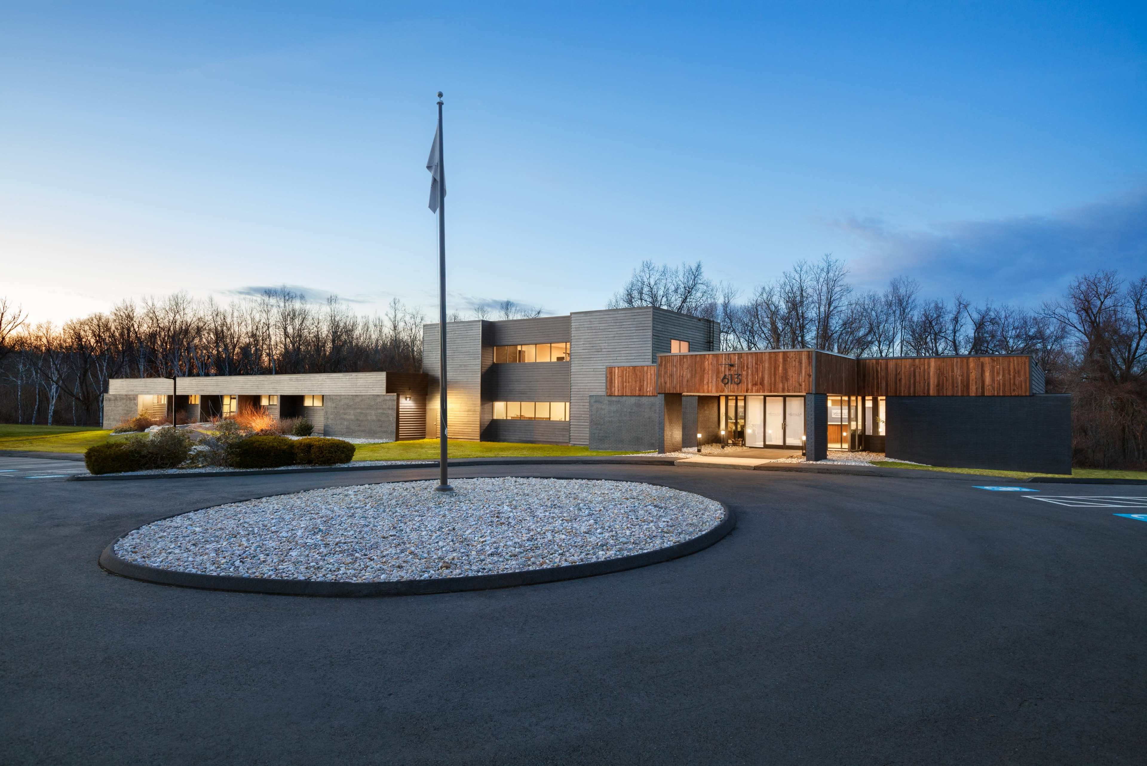 A modern building with a sleek facade and a circular gravel driveway is situated in a landscaped area surrounded by trees.