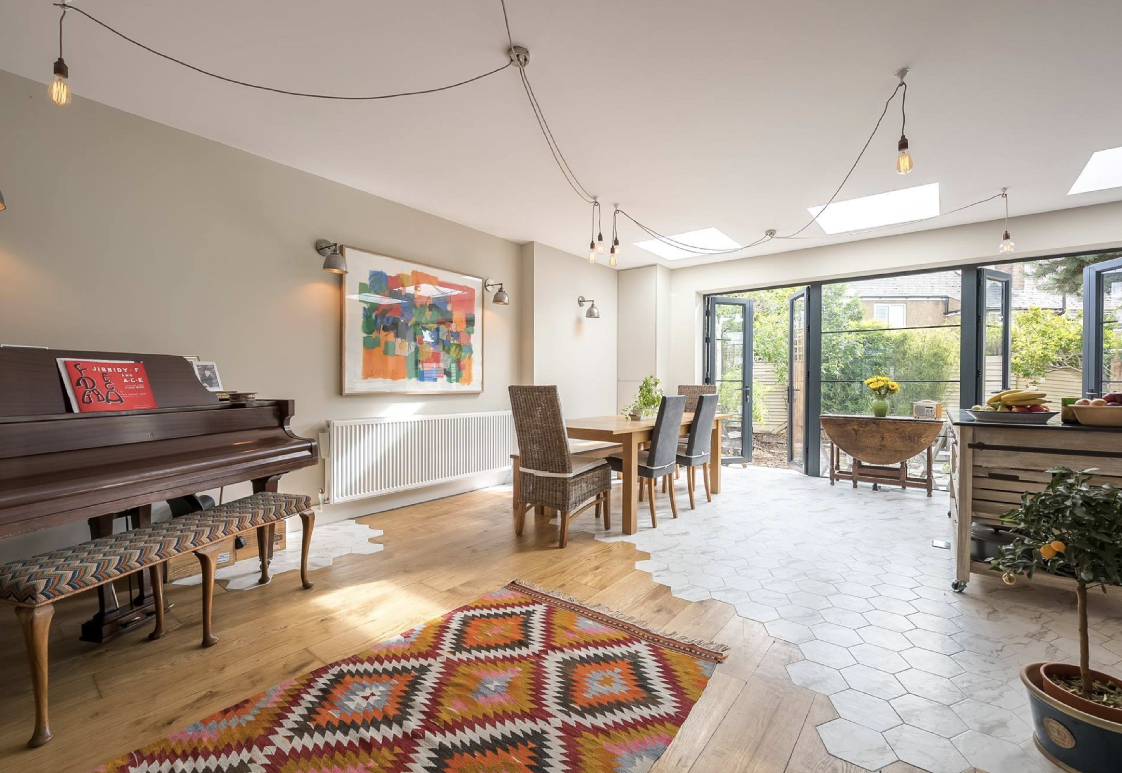 The image shows a bright dining area with a piano, a wooden dining table and chairs, and large glass doors leading to a garden.