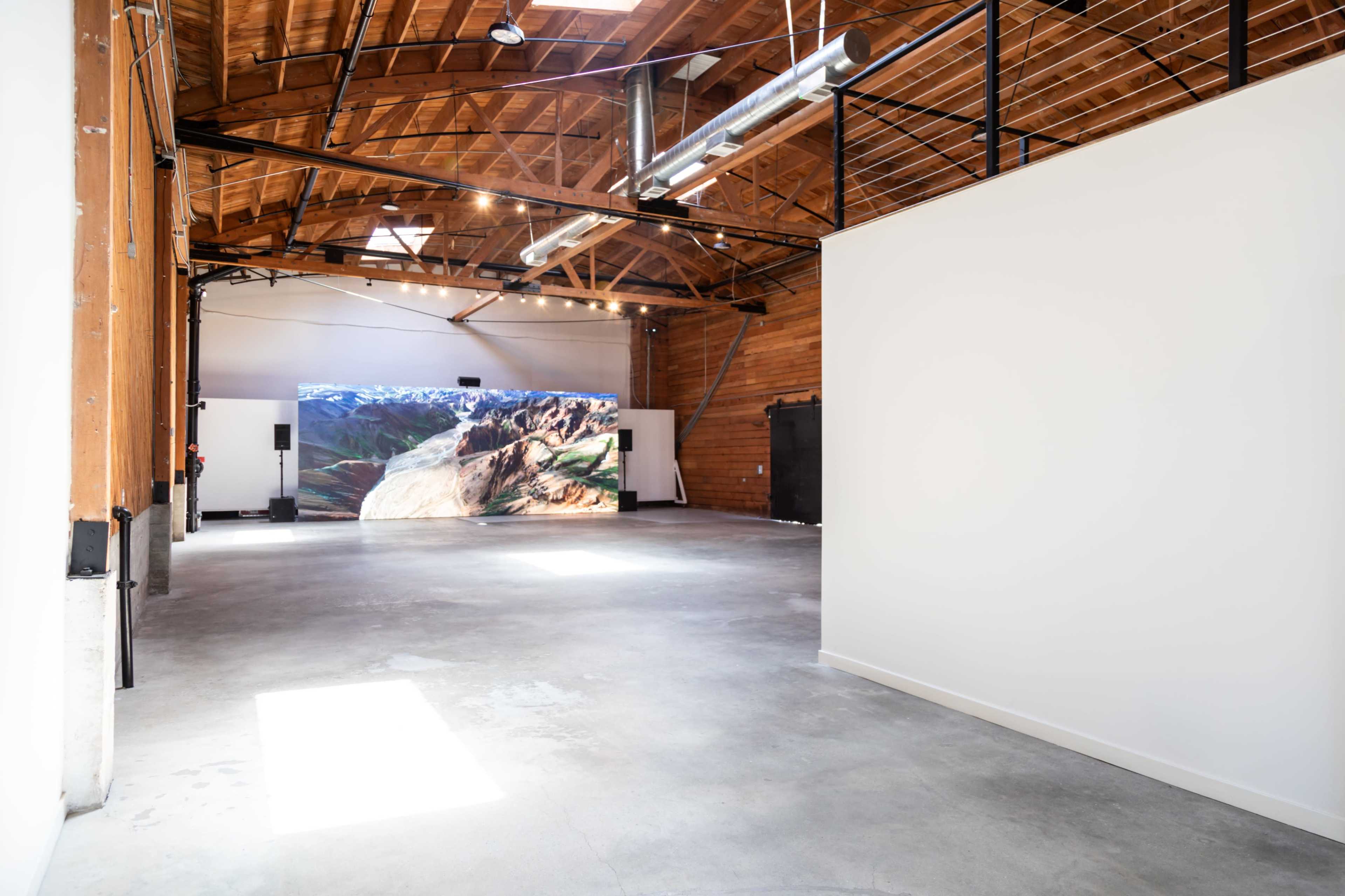 A spacious, open gallery with a large video projection displaying a landscape on one wall.