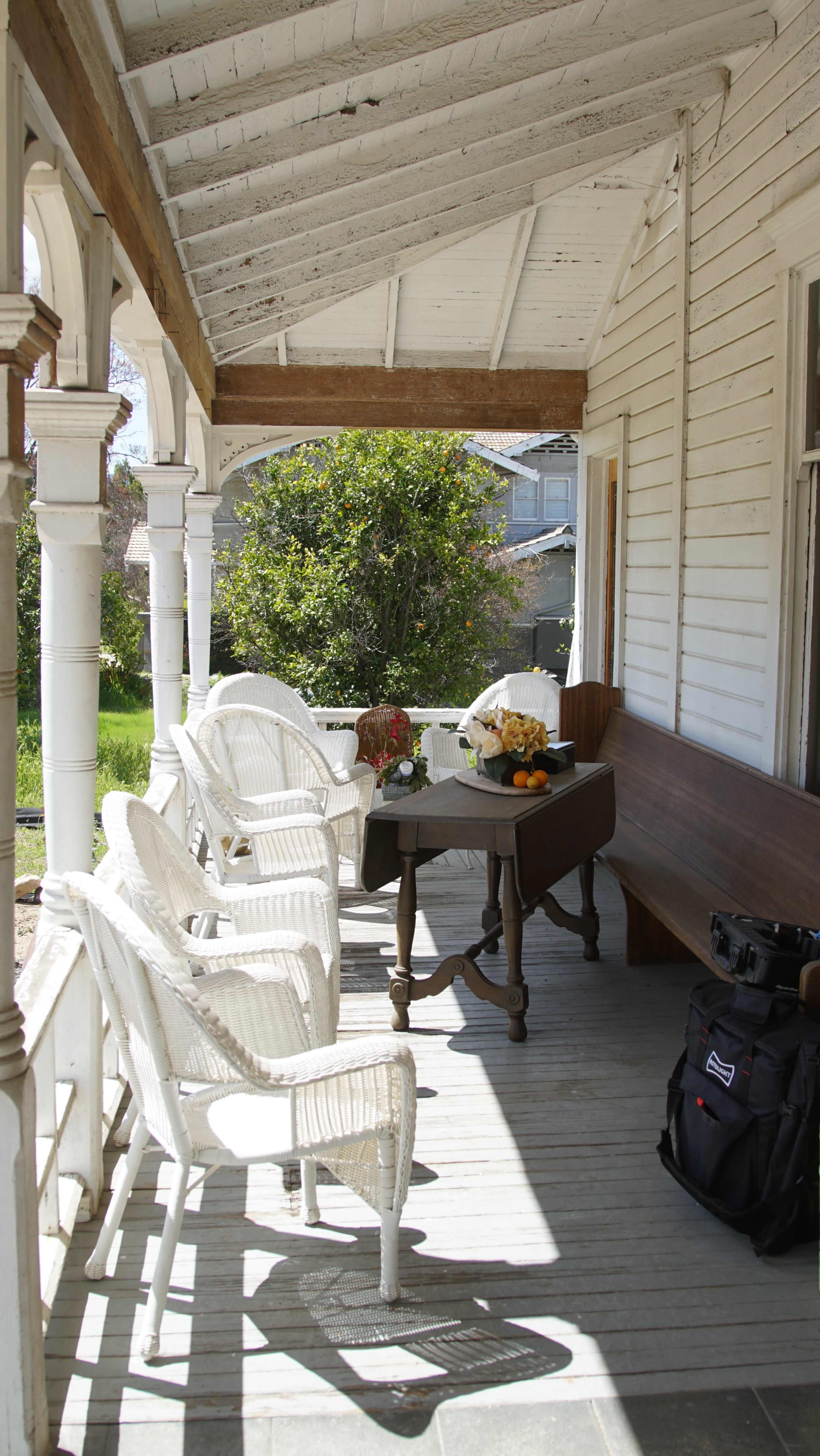 A porch features wicker chairs arranged around a table adorned with flowers and fruit, with a bag resting nearby.