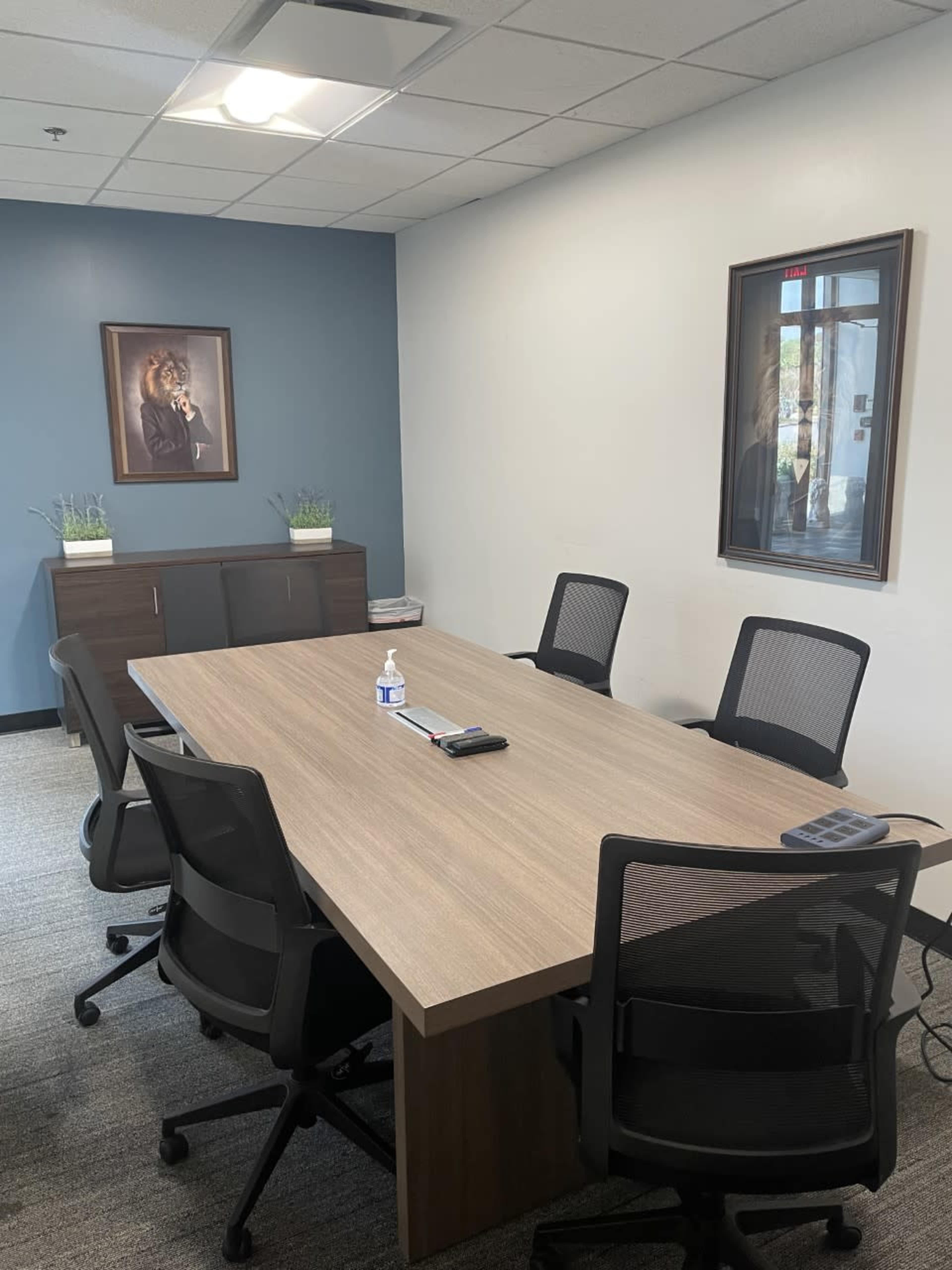 A conference room features a large wooden table surrounded by six chairs, with artwork on the walls and minimal decorations.