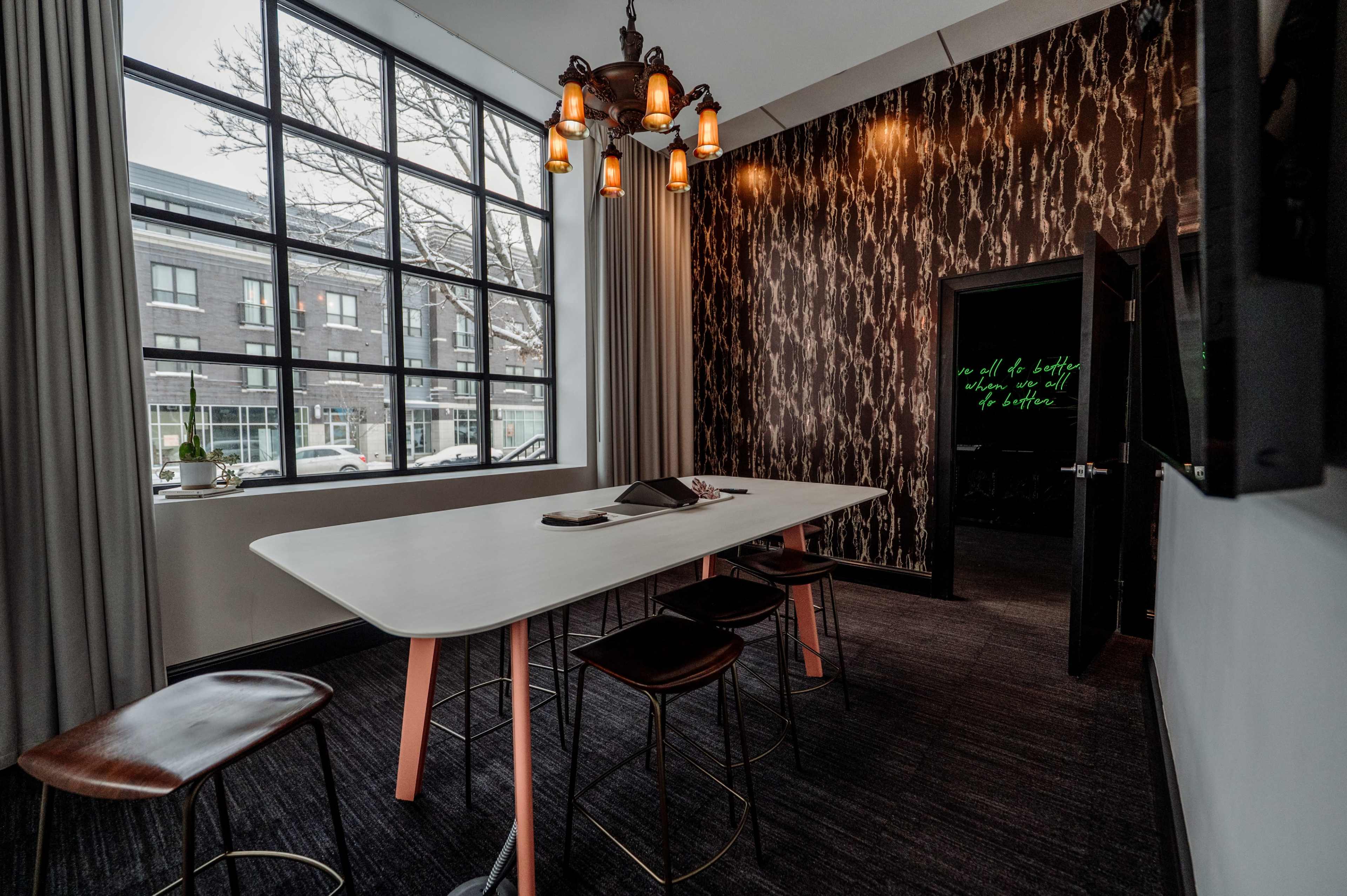 The image shows a modern meeting room with a long white table, four wooden stools, and a wall adorned with dark patterned wallpaper, illuminated by a chandelier with multiple bulbs.