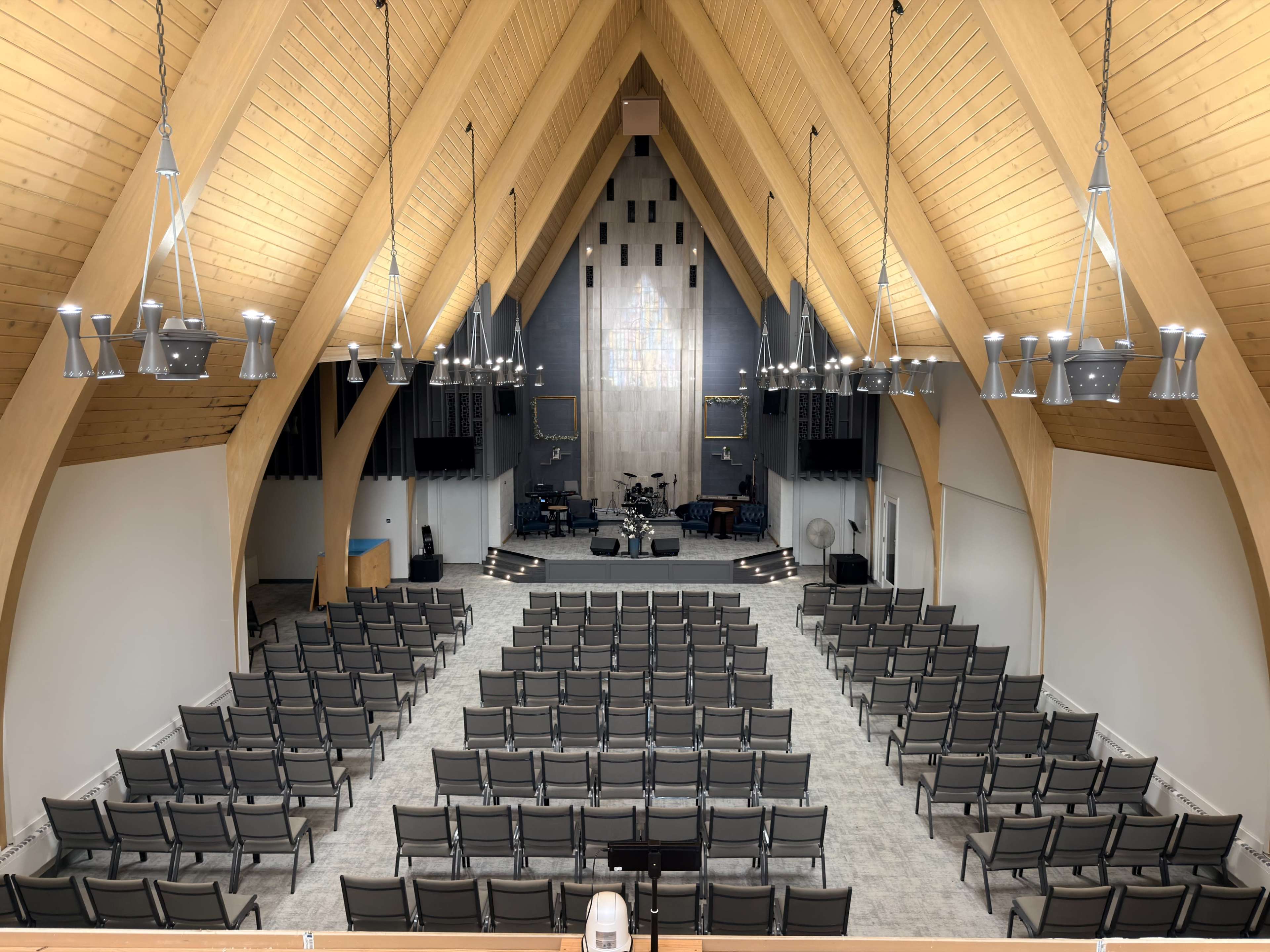 The image shows an interior view of a spacious church auditorium with wooden beams, rows of chairs, and a stage equipped with musical instruments.
