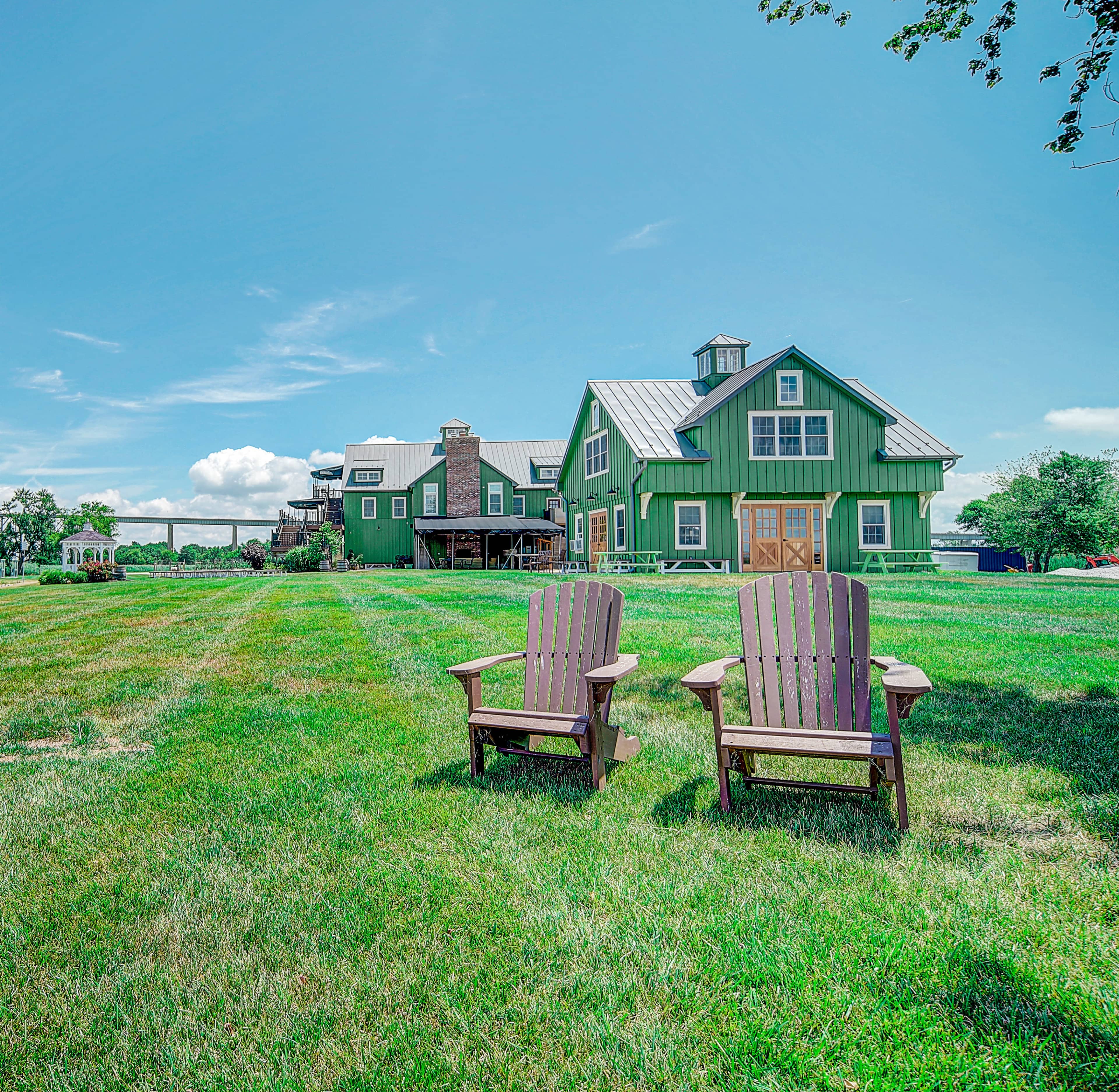 Two wooden chairs sit in a grassy area in front of a green farmhouse under a clear blue sky.
