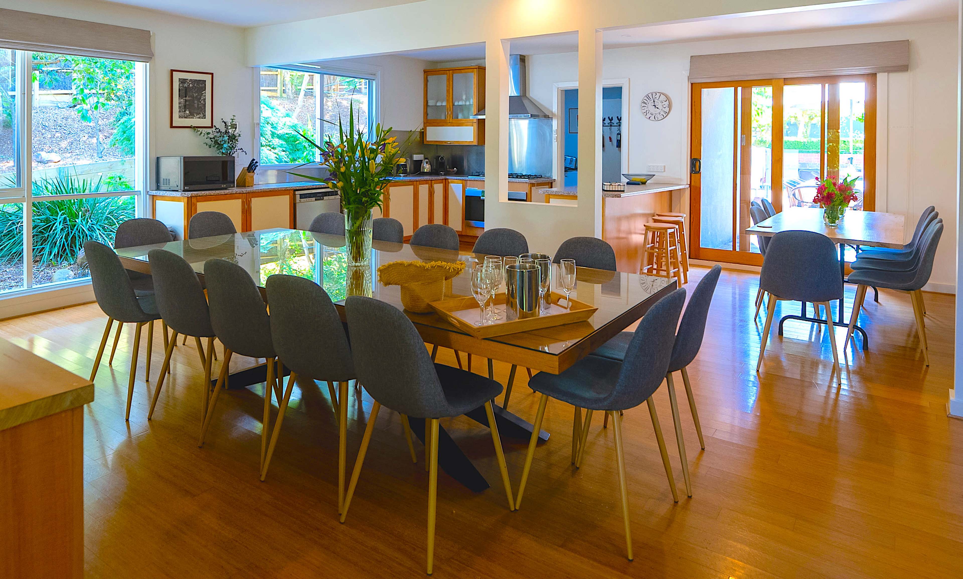 A spacious dining area with a large table surrounded by gray chairs, flanked by a kitchen and sliding glass doors leading to an outdoor space.