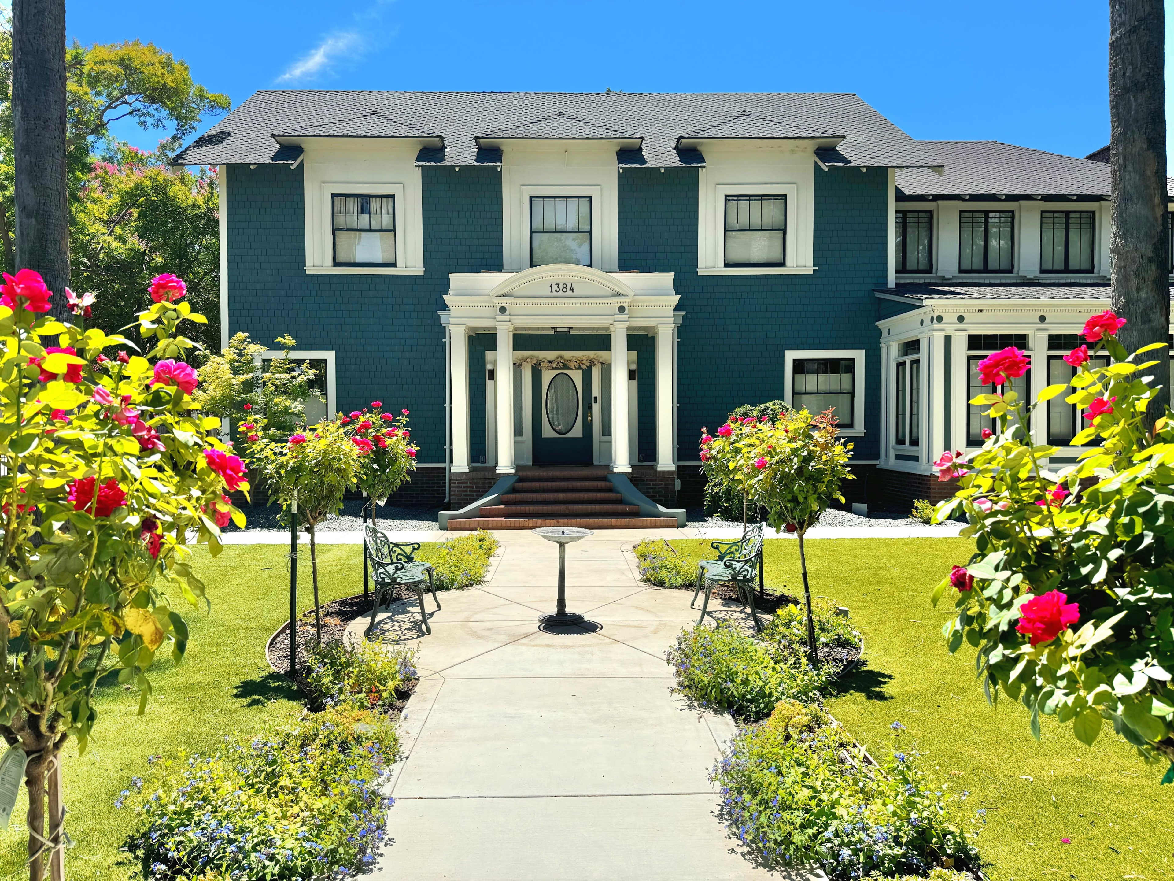 A large, two-story blue house with white trim is framed by well-maintained gardens featuring blooming rose bushes and a central pathway leading to the front entrance.