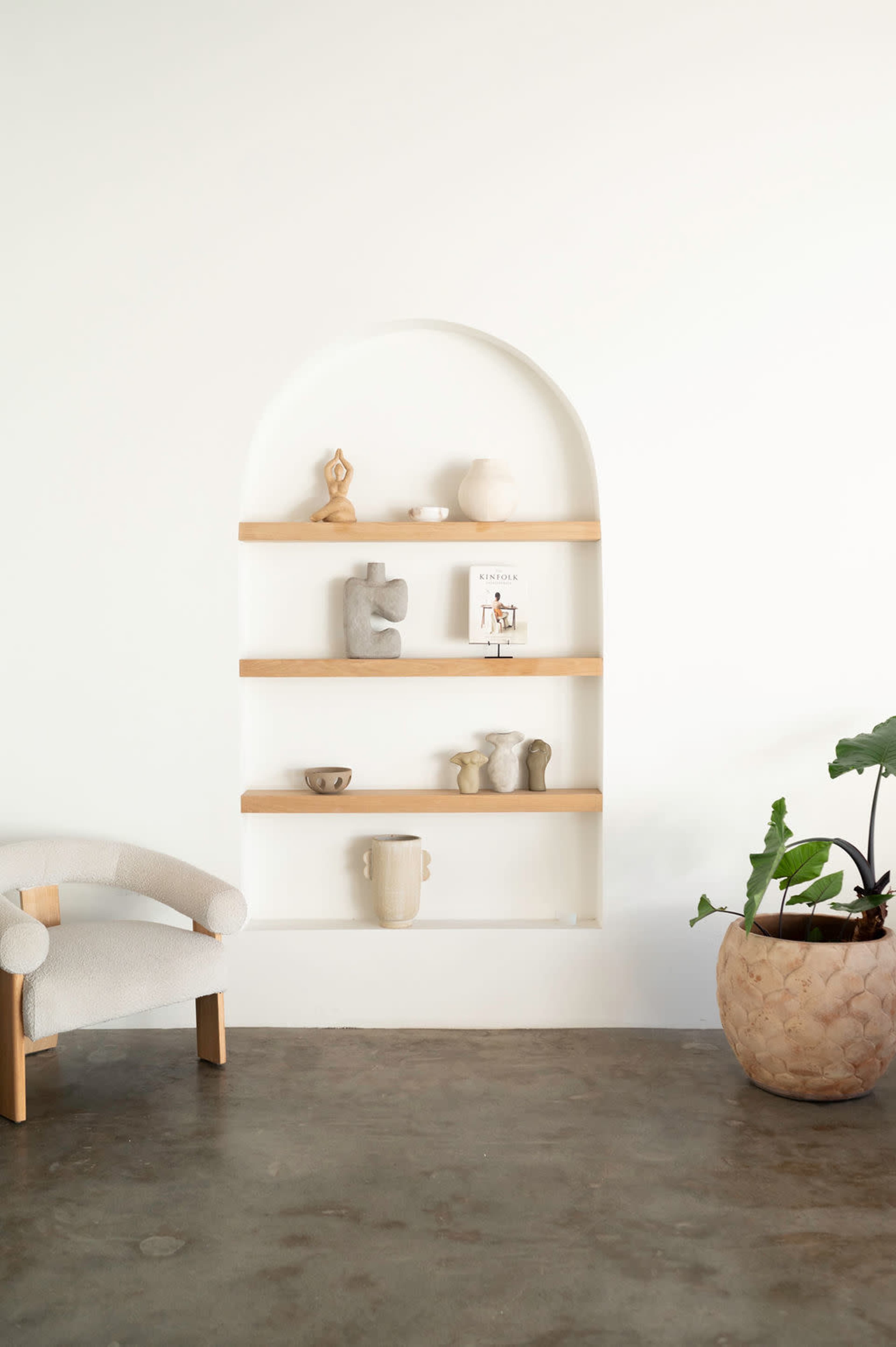 A minimalist room features a curved wall niche with wooden shelves displaying various decorative objects and a small plant beside a light-colored upholstered chair.