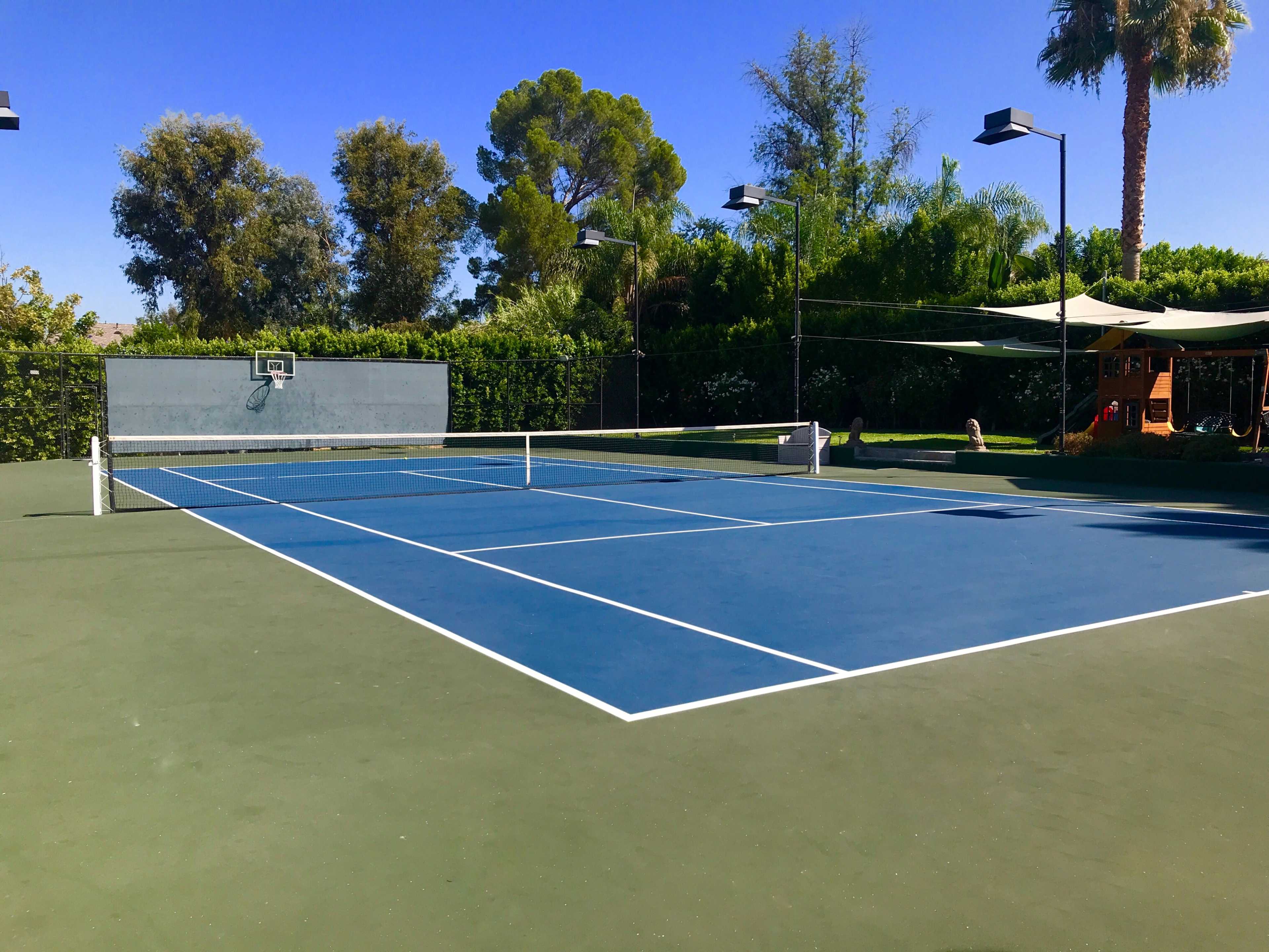 A well-maintained tennis court with blue lines and a hoop in the background, surrounded by greenery and palm trees.