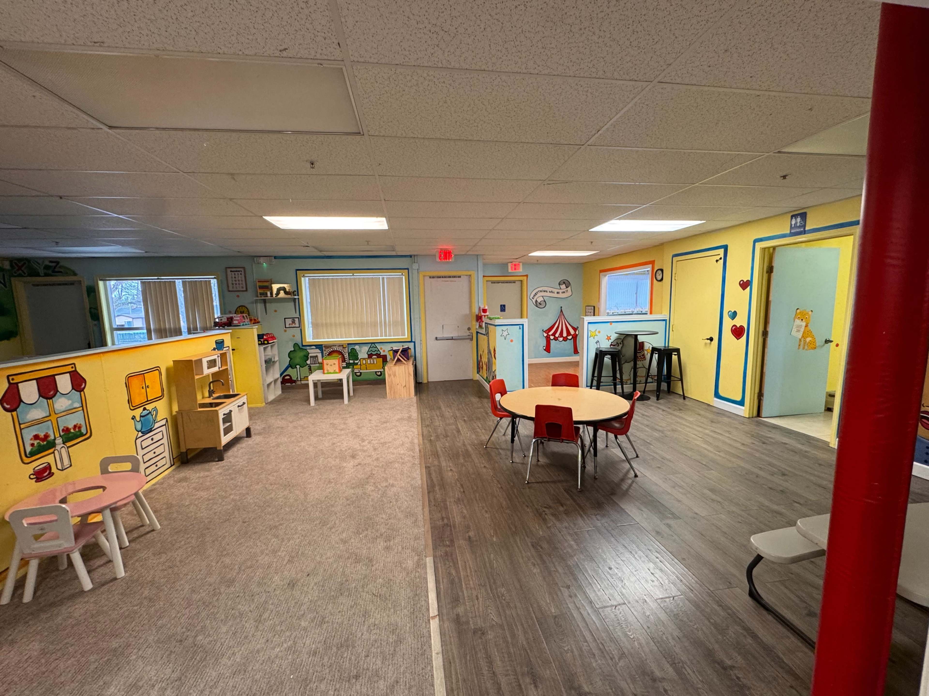 The image shows a brightly colored classroom divided into two areas, one with small tables and chairs and the other featuring play equipment and storage.