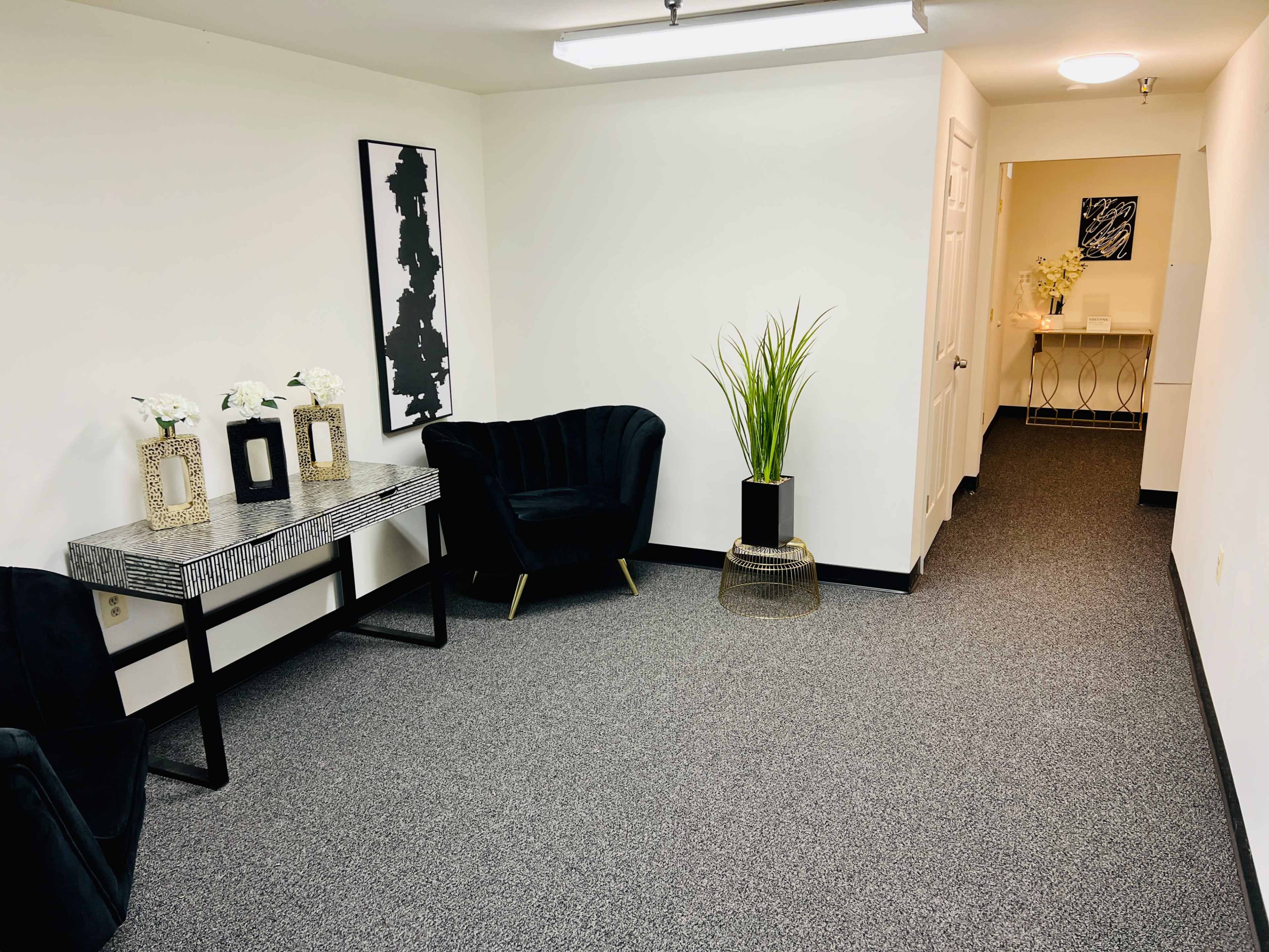 A small waiting area with a black sofa, a decorative table with flower arrangements, a tall potted plant, and a hallway leading to another room.