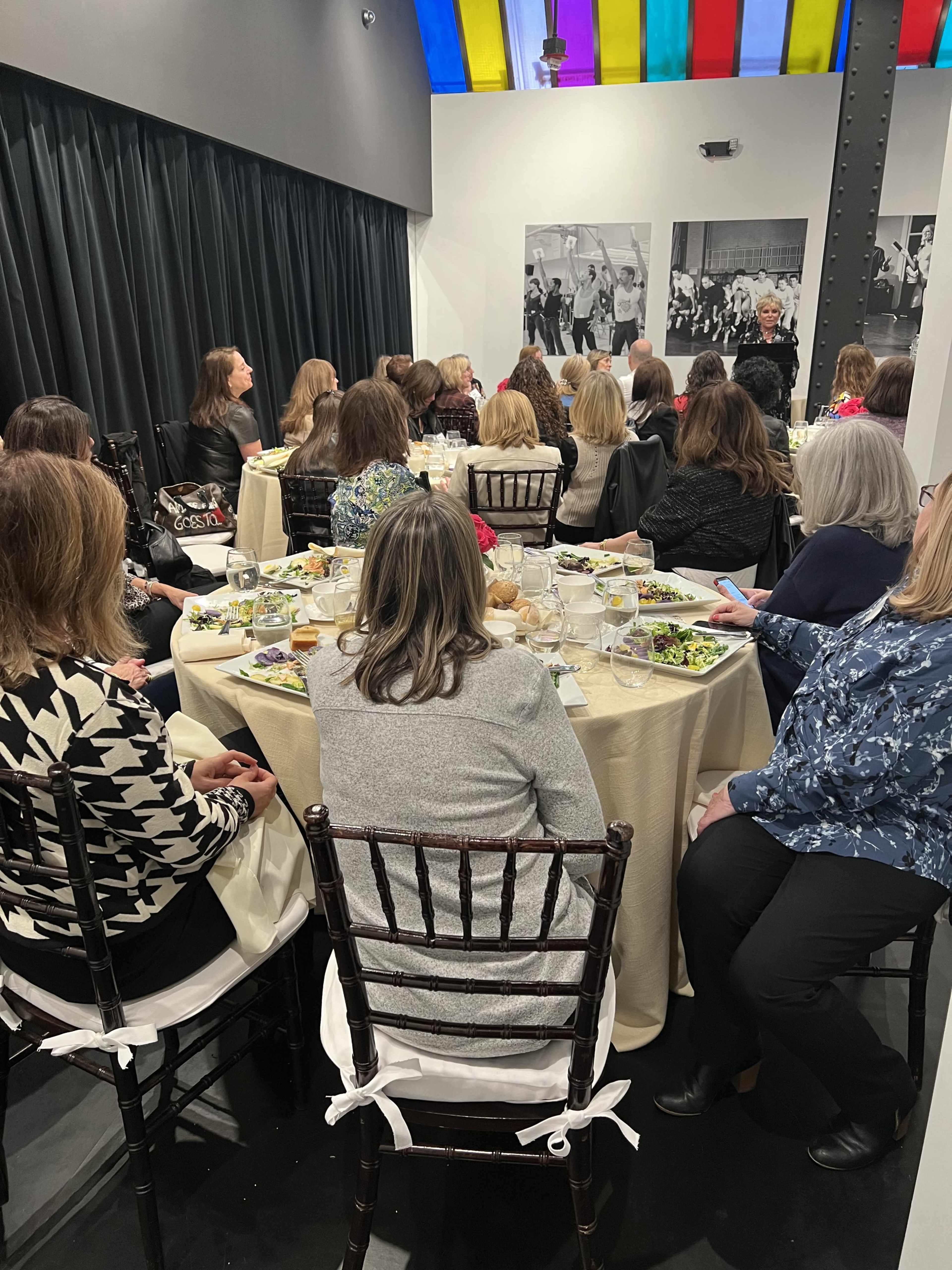 A group of women sits at tables covered with white linens in a well-lit room, listening to a speaker in front of black-and-white photographs on the walls.