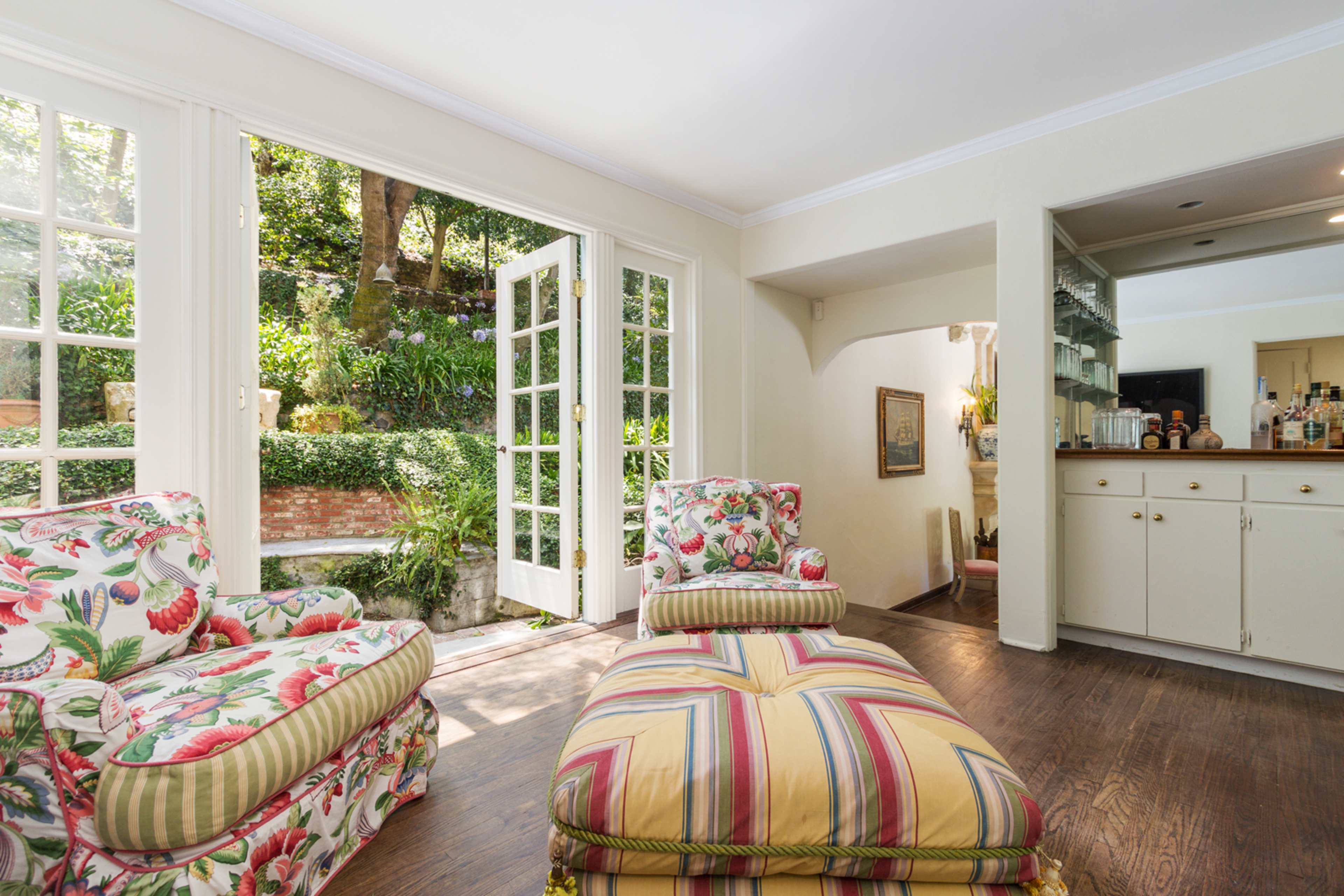 The image depicts a bright living room with floral-patterned armchairs and a view of a lush garden through glass-paneled doors.