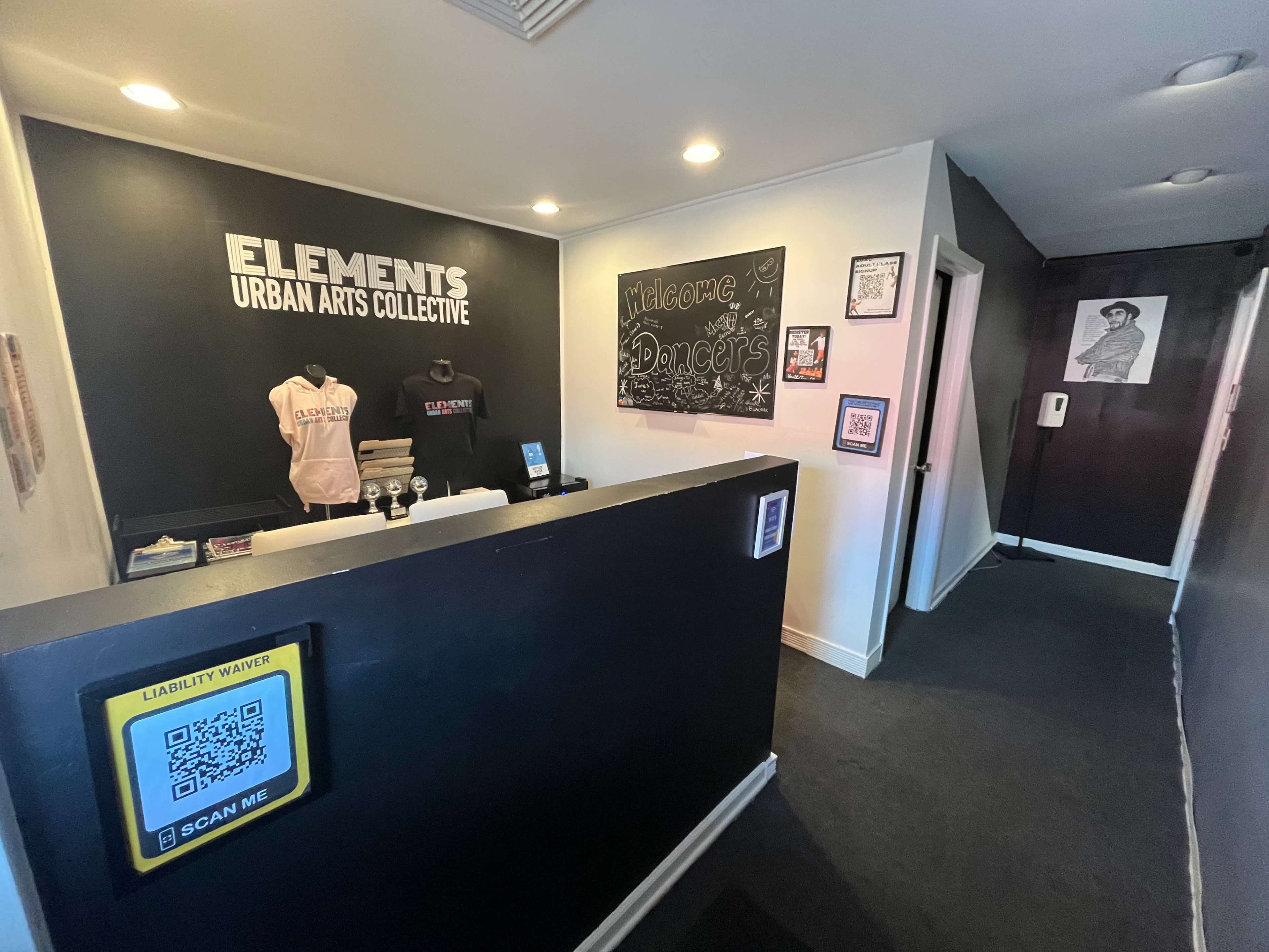 A reception area for "Elements Urban Arts Collective," featuring a black wall with signage, merchandise on display, and a hallway leading to another room.