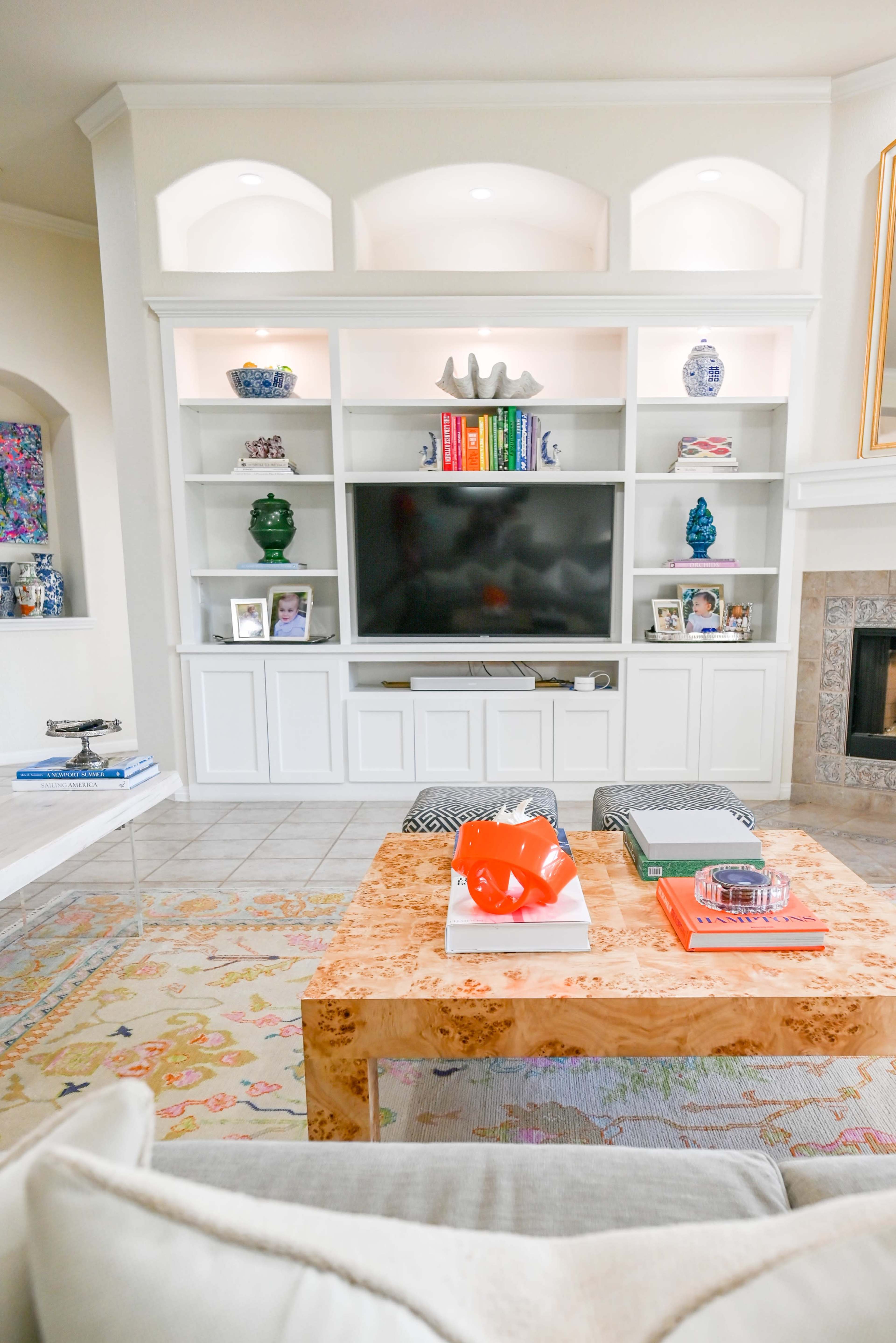A modern living room features a built-in entertainment center with shelves displaying decorative items and a large television, complemented by a central coffee table and a patterned area rug.