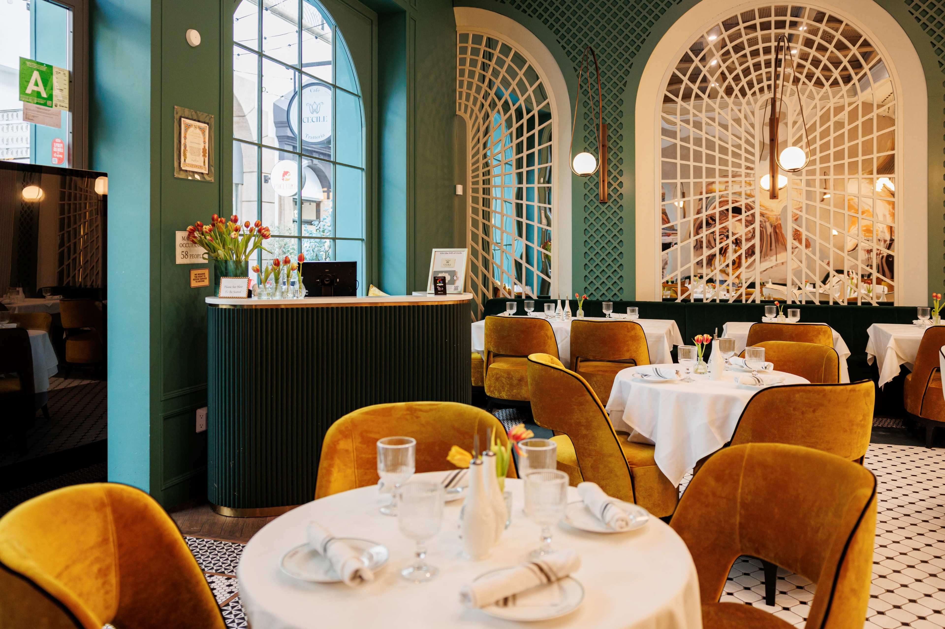 The image shows a beautifully arranged restaurant interior with round tables, yellow upholstered chairs, and decorative green walls featuring large windows and architectural details.