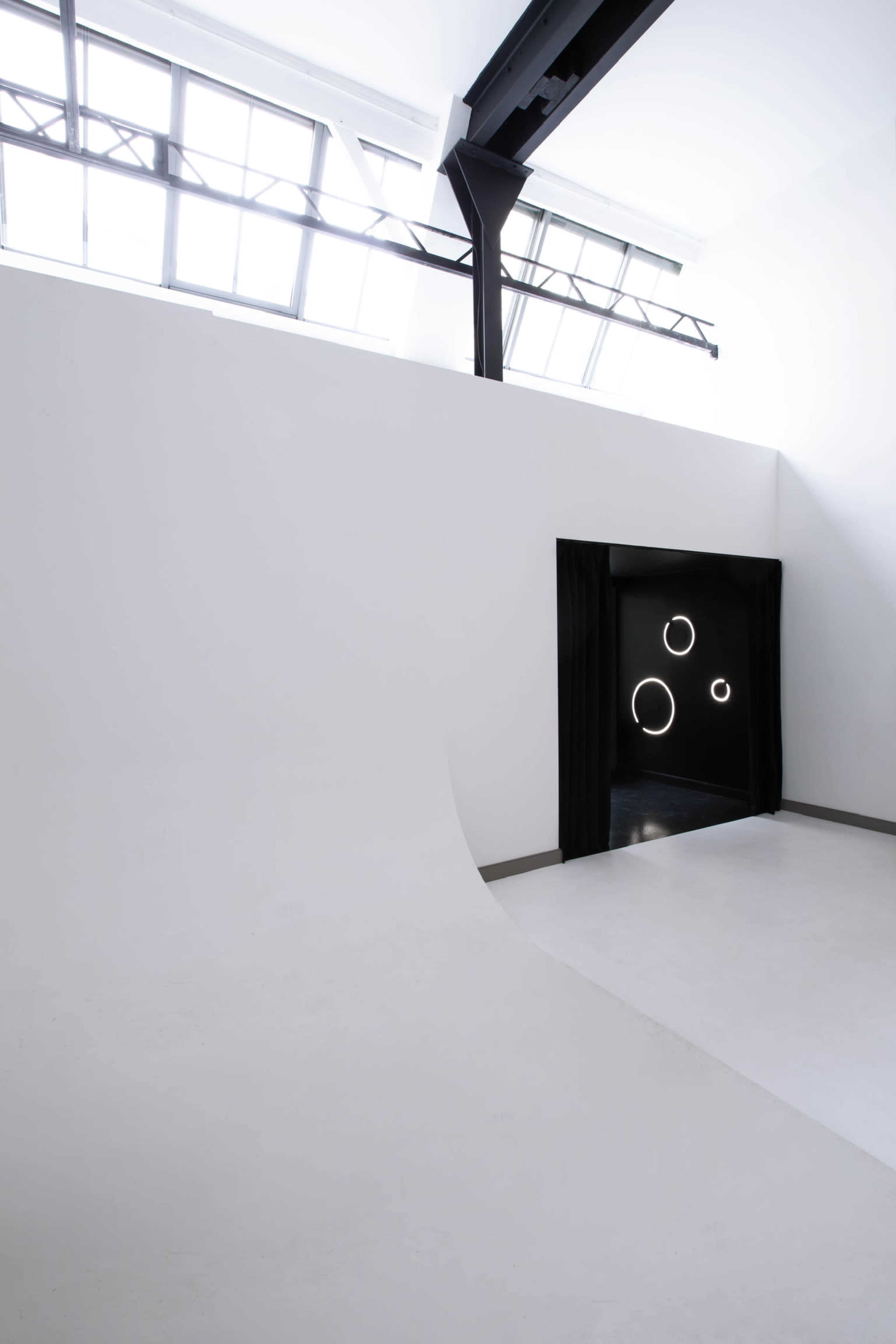 The image shows a modern, minimalistic interior space with a curved white wall leading to a dark doorway, illuminated by circular lights.