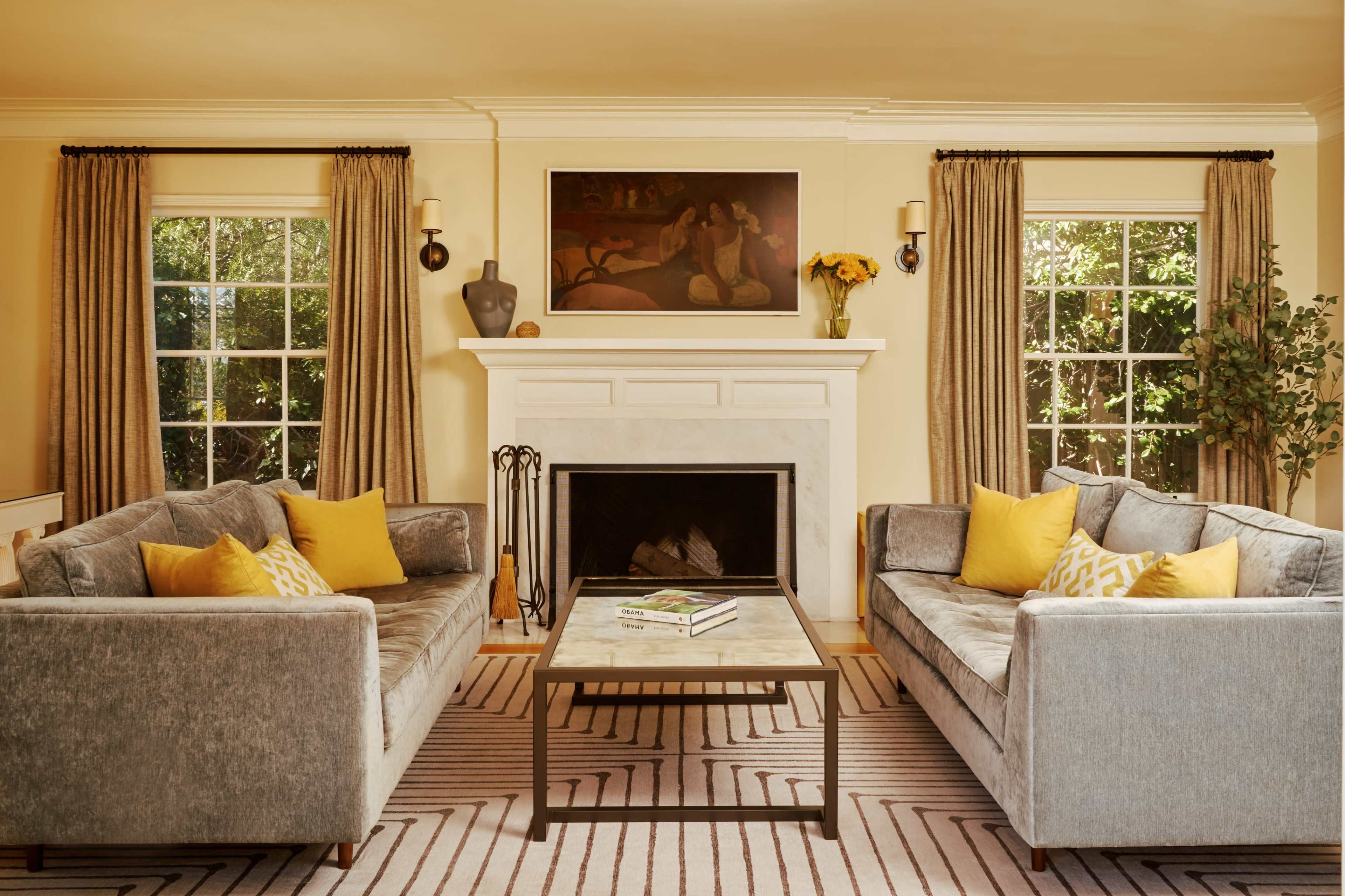 The living room features two gray sofas with yellow accent pillows arranged around a coffee table in front of a fireplace adorned with a painting and flanked by windowed walls.
