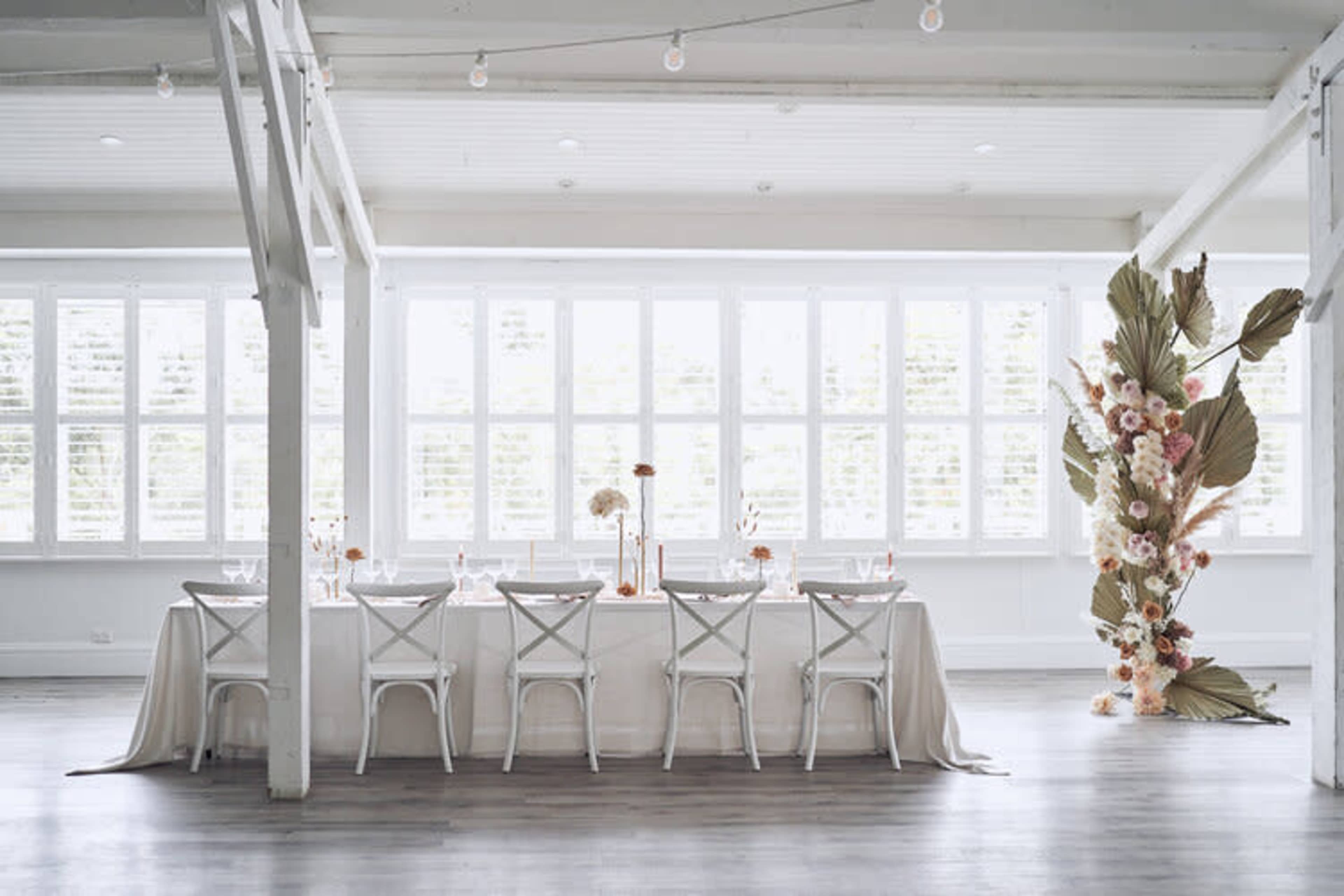 A long table set for dining is positioned in a brightly lit room with large windows, accompanied by a floral arrangement at one end.