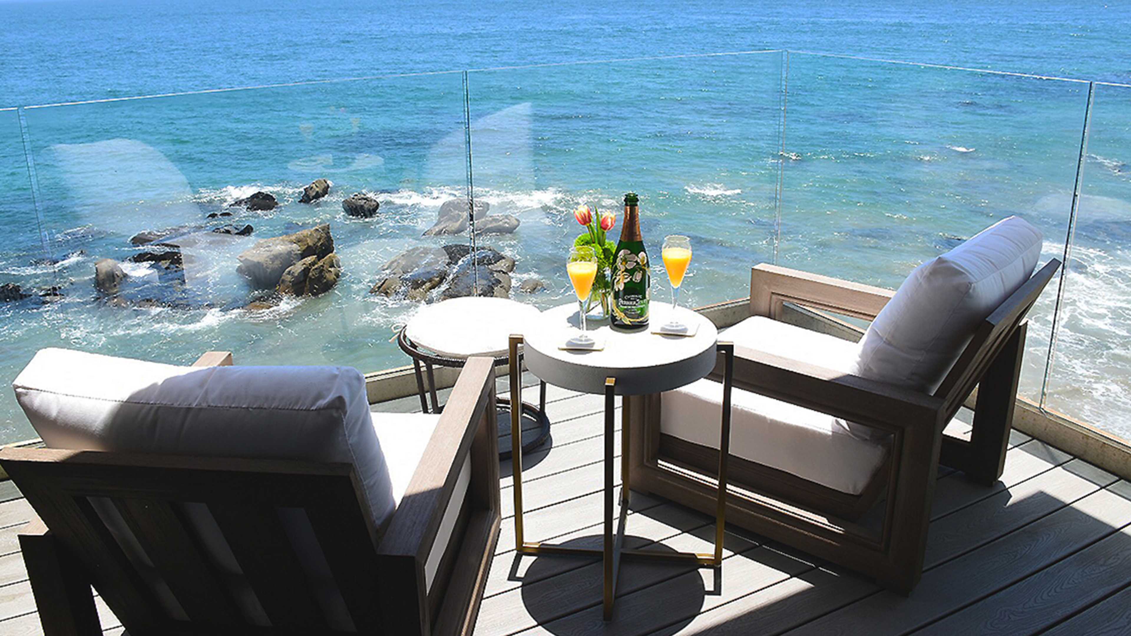A balcony overlooking the ocean features two wooden chairs, a small table with a bottle of champagne and two drinks, and a clear glass railing.
